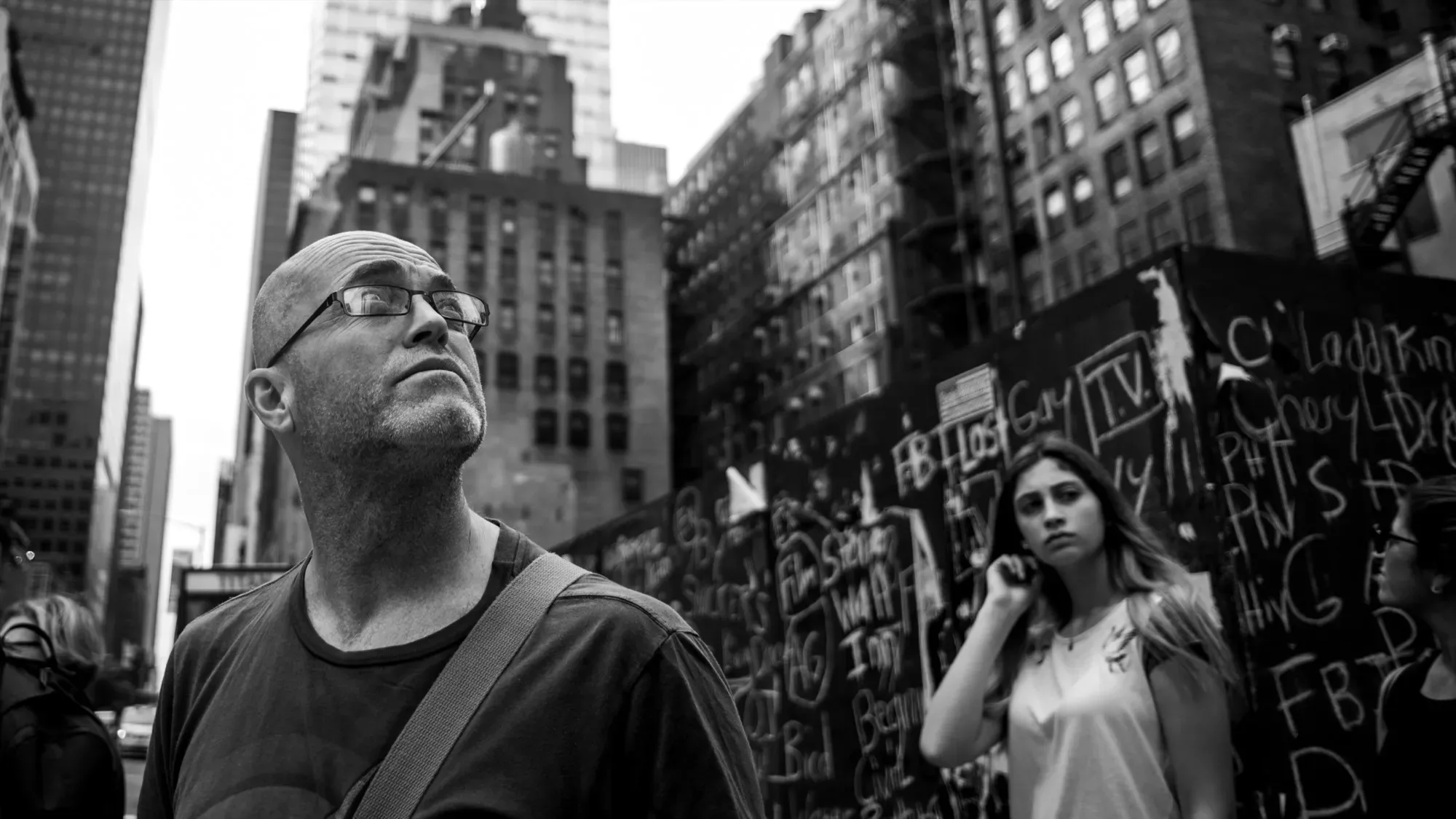 Black-and-white photo of Paul Kenton gazing up at tall buildings on a busy New York street, a chalk-covered hoarding behind him
