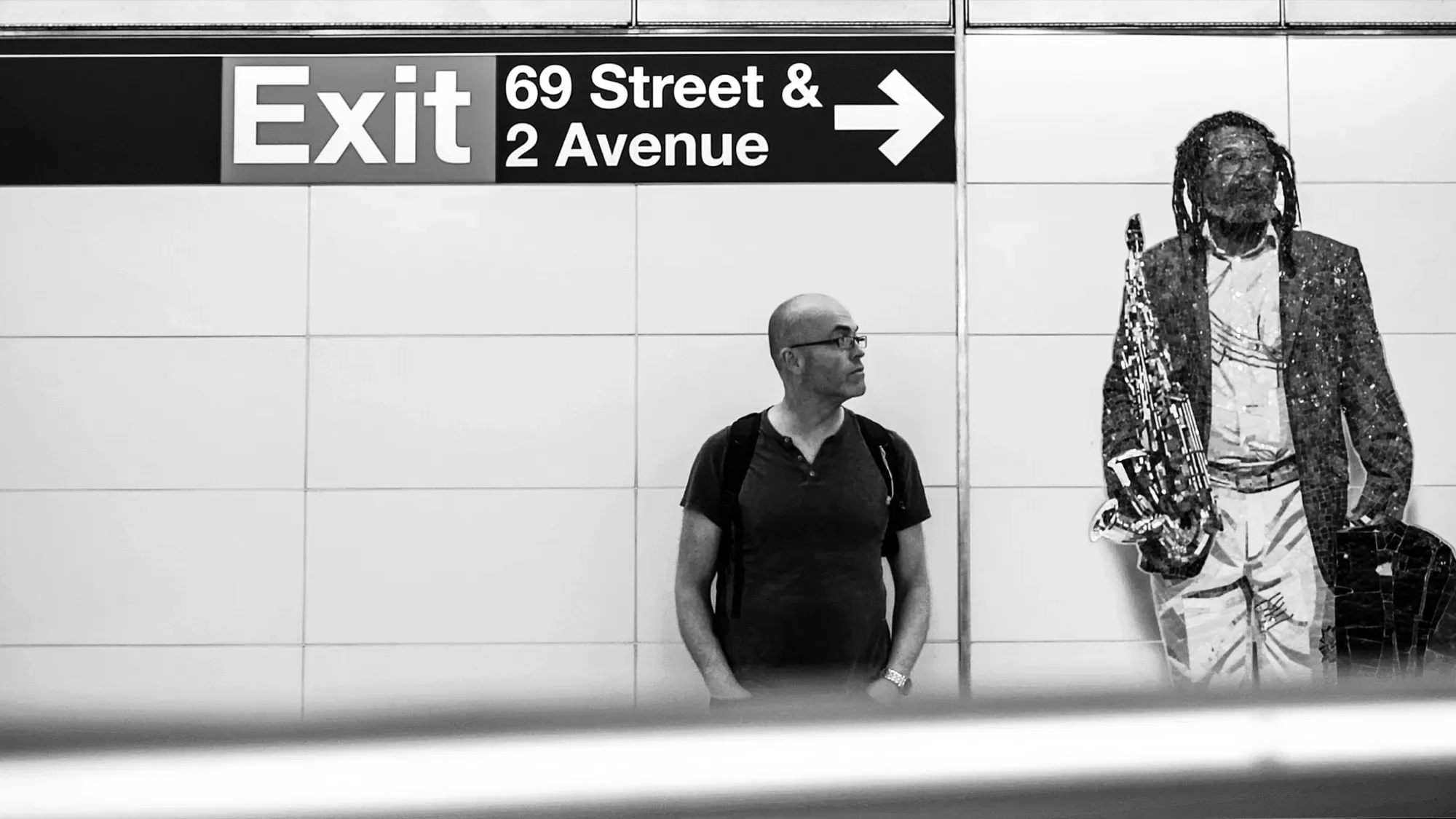 Black-and-white photo of Paul Kenton on a New York subway platform under an Exit 69 Street & 2 Avenue sign, beside a mosaic of a saxophonist