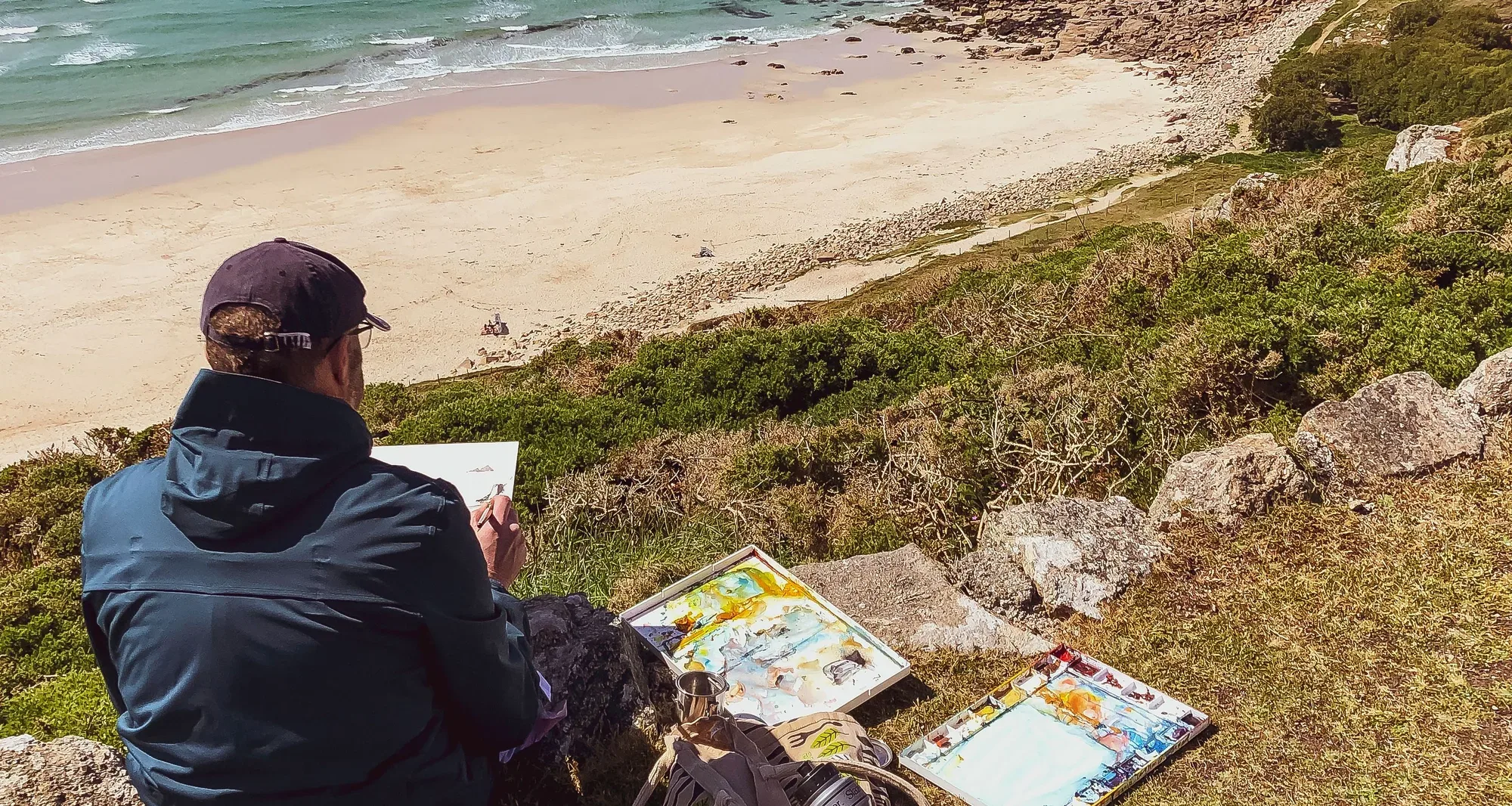Paul Kenton seen from behind, sitting on a grassy clifftop painting in watercolour, finished sketches laid out beside him above a beach and turquoise sea
