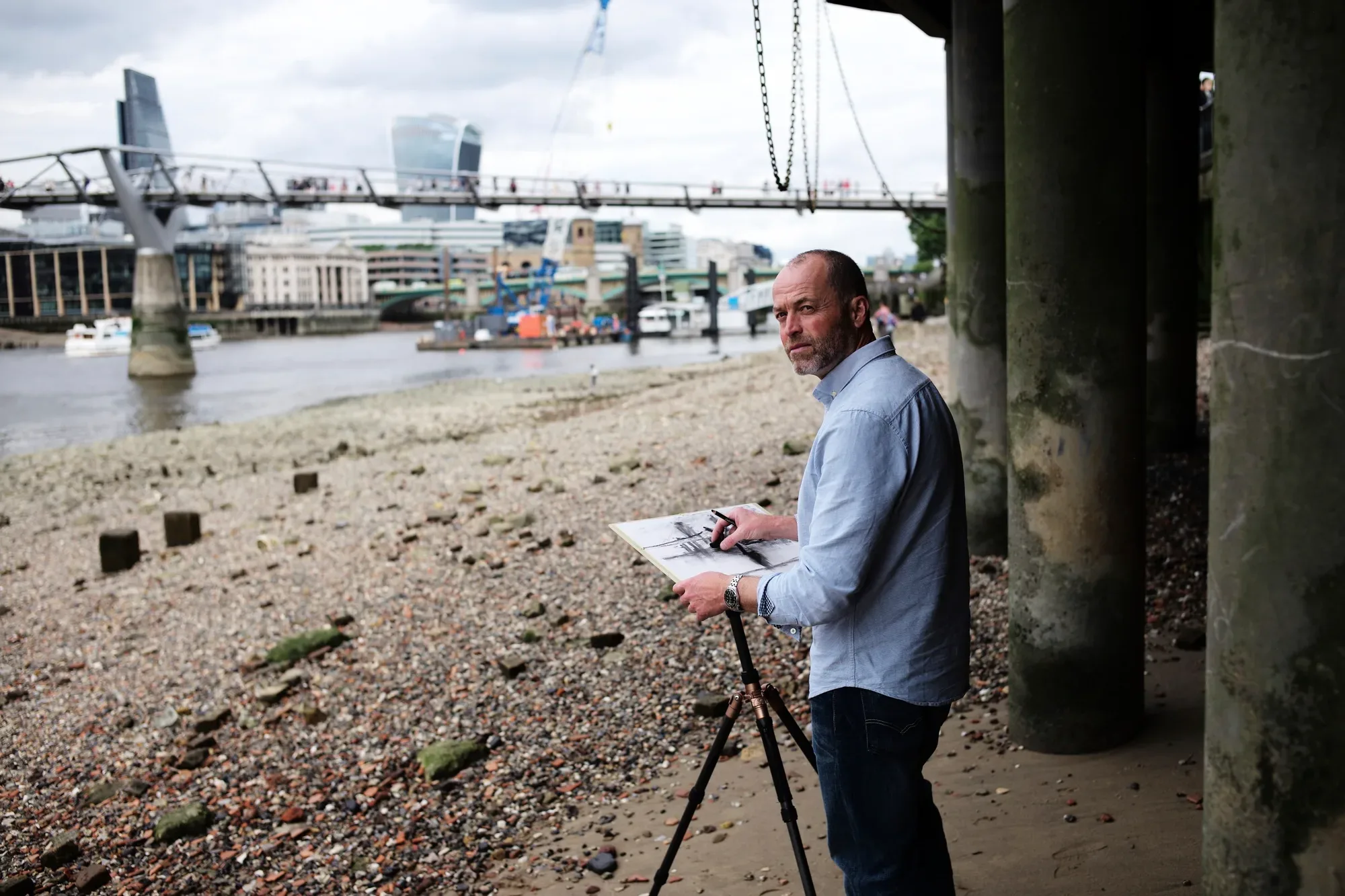 Paul Kenton sketching at an easel on the shingle of the Thames foreshore, the Millennium Bridge and City of London towers behind him