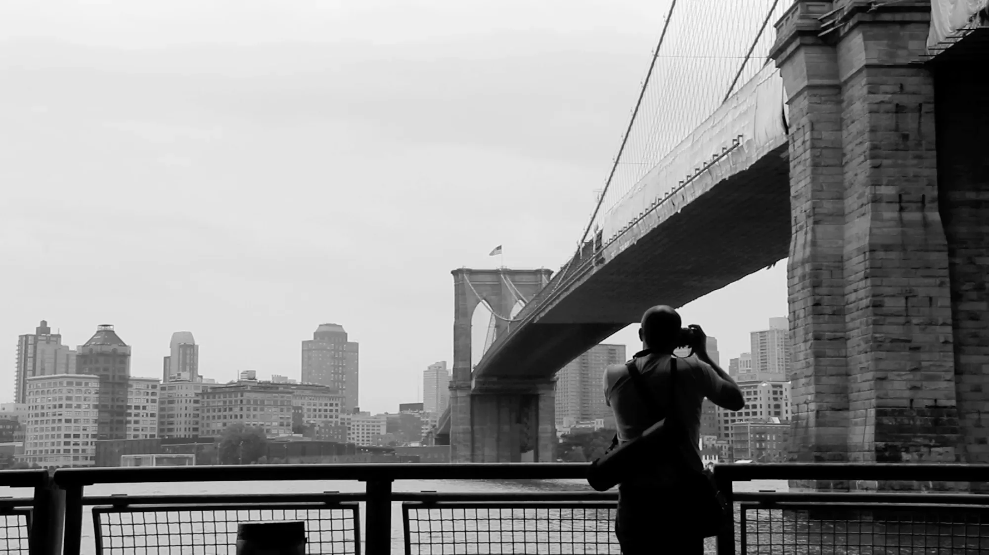 Black-and-white photo of Paul Kenton from behind, photographing the Brooklyn Bridge from a railing on the waterfront