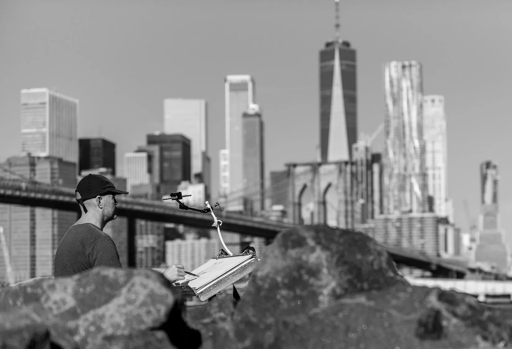 Black-and-white photo of Paul Kenton sketching at an easel among rocks, with One World Trade Center and the Manhattan skyline behind him