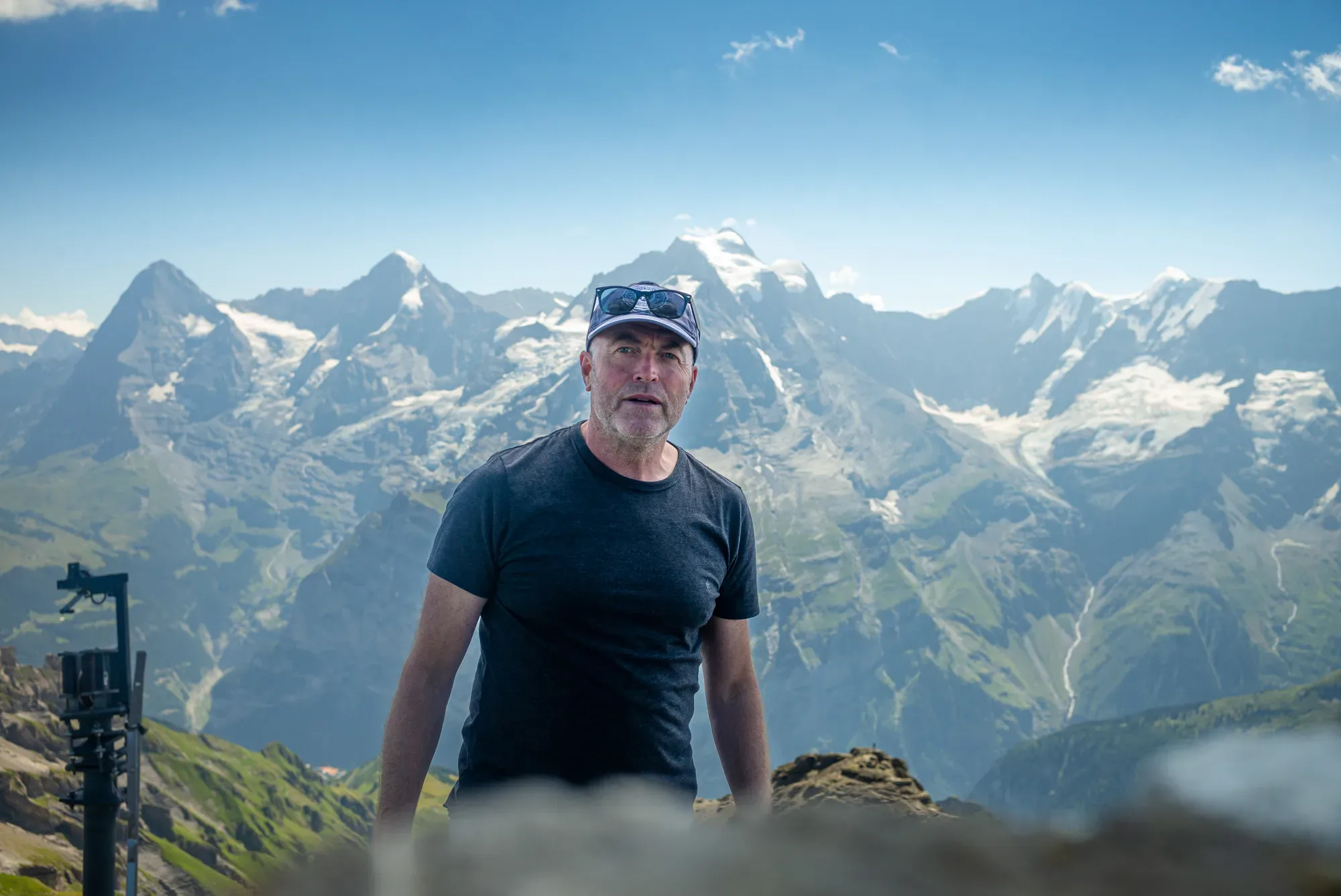 Paul Kenton in a cap and sunglasses standing in front of a range of snow-capped Alpine peaks