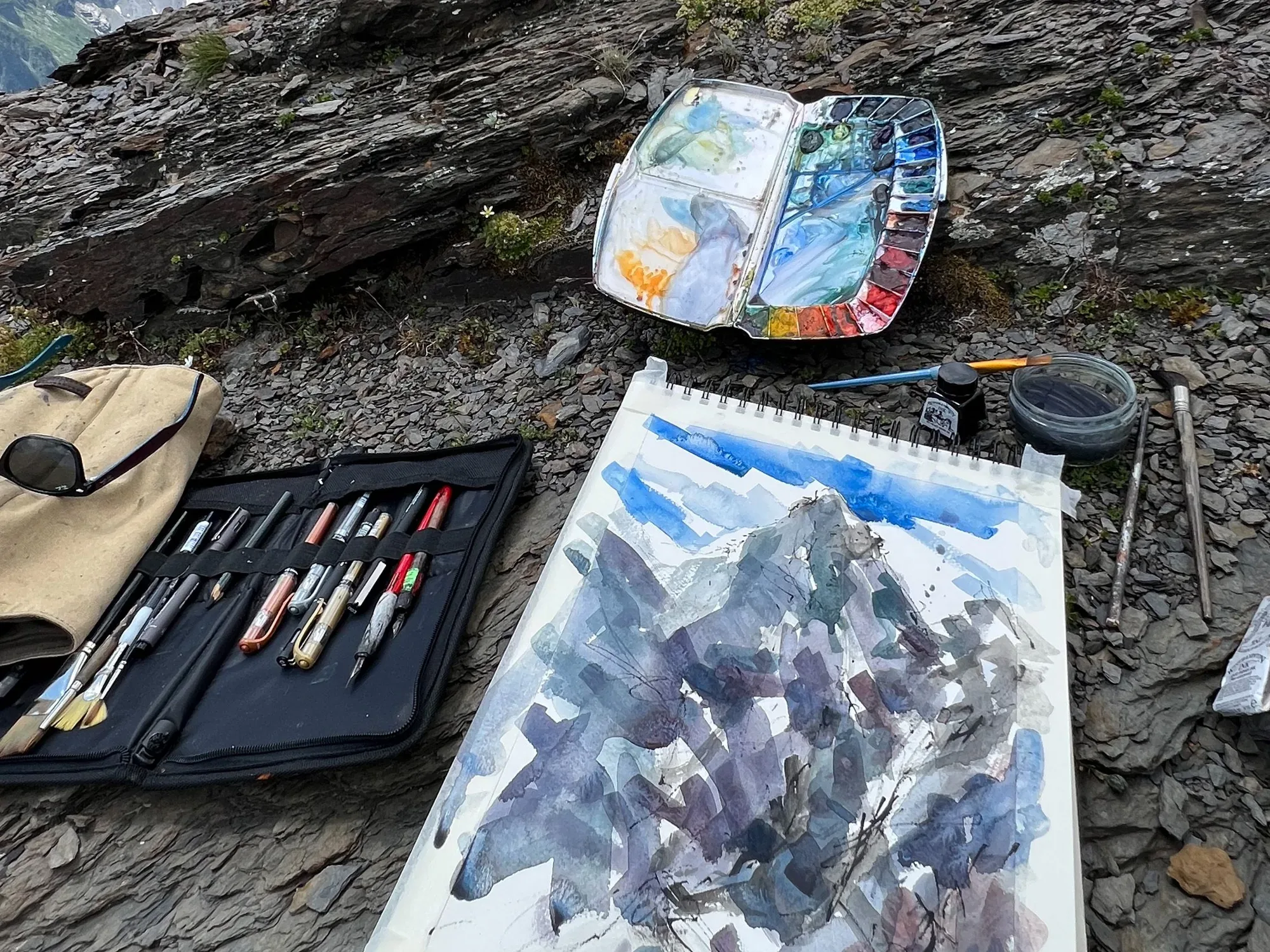 A watercolour sketch of a mountain in blues and purples on a pad, with a paintbox and a case of pens laid out on slate rock