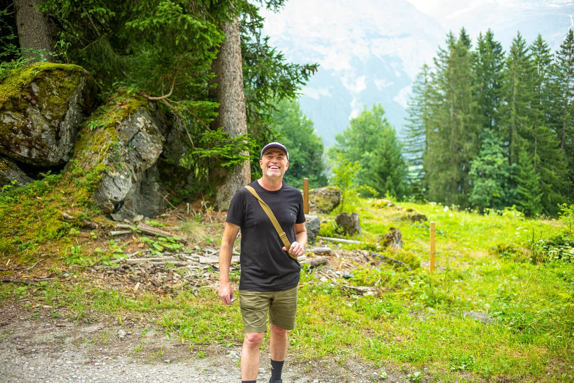 Paul Kenton smiling on a woodland path in the Swiss mountains, pine trees and a misty peak behind him