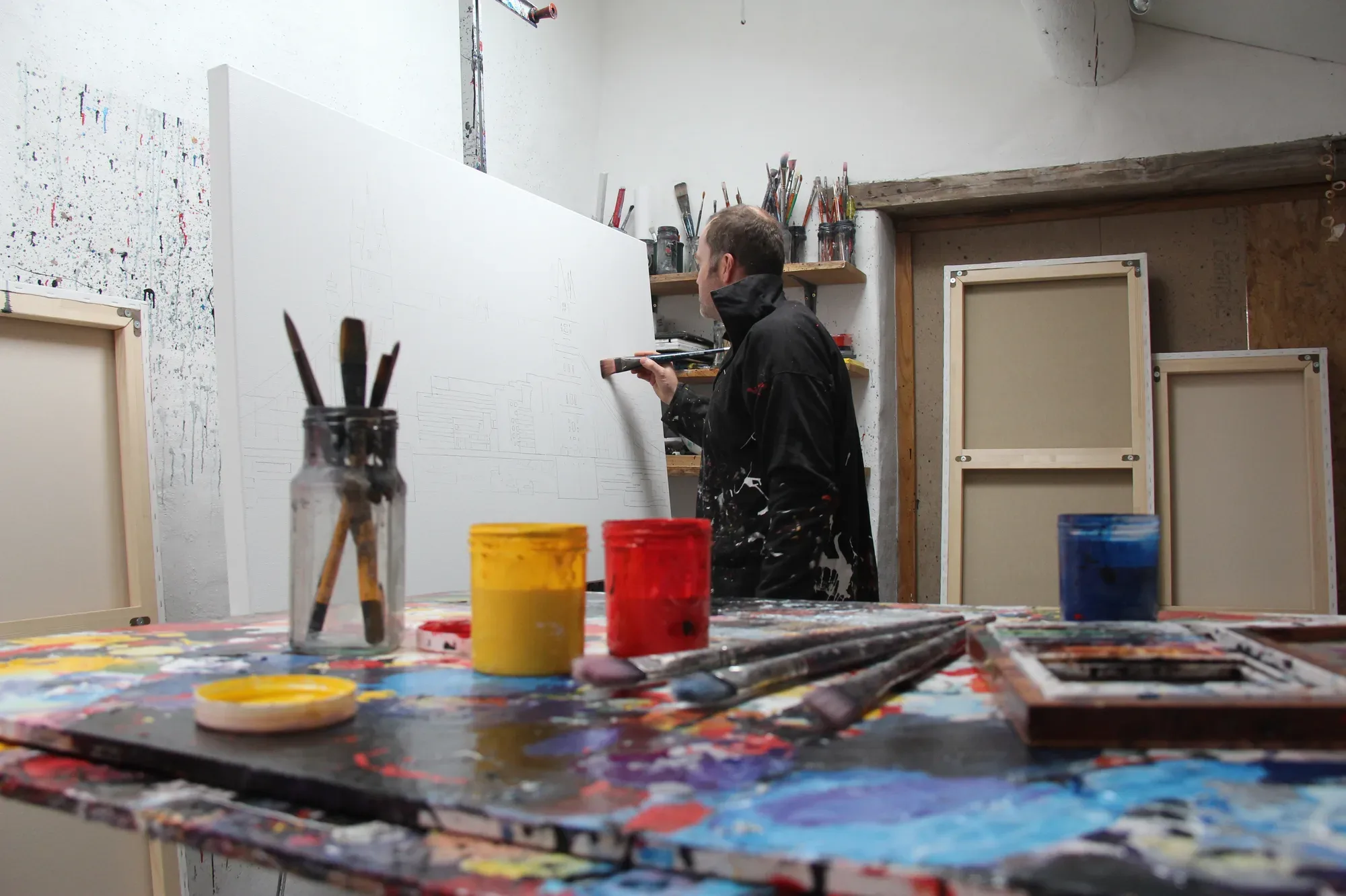 Paul Kenton in a paint-covered jacket drawing outlines on a large white canvas, pots of yellow and red paint on the table in front