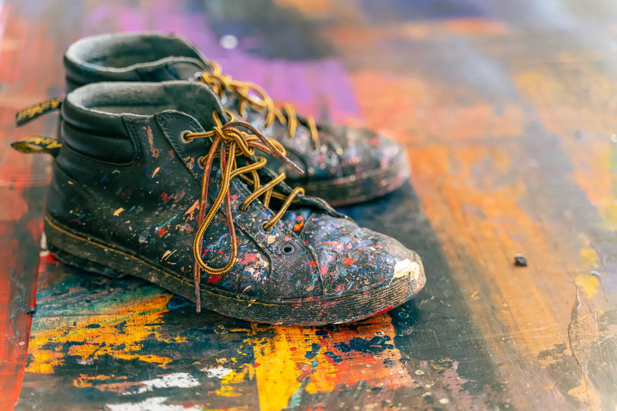 A pair of black boots spattered with paint standing on a floor covered in layers of coloured paint