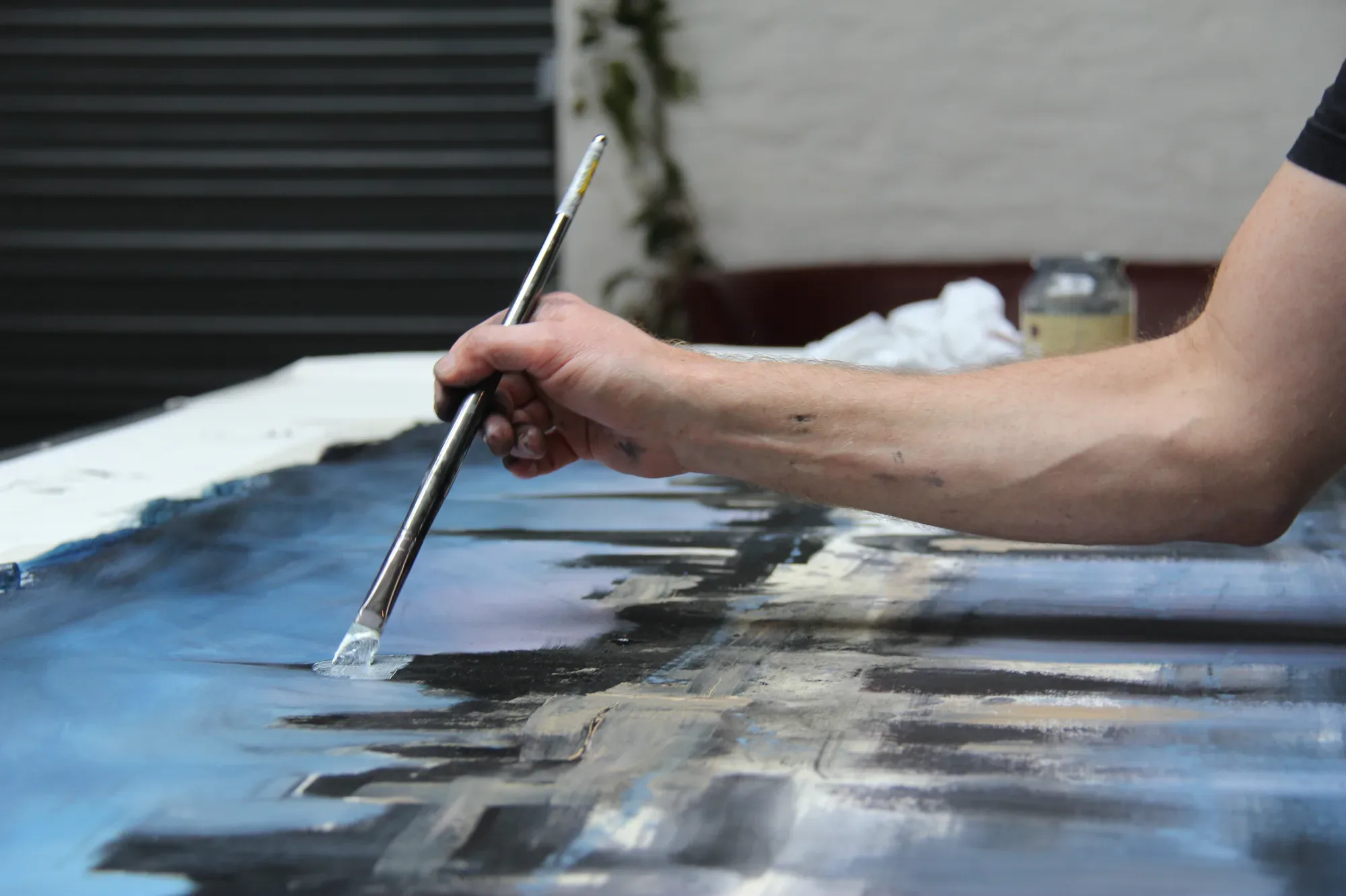 Close-up of a hand holding a long brush against a blue and black cityscape canvas