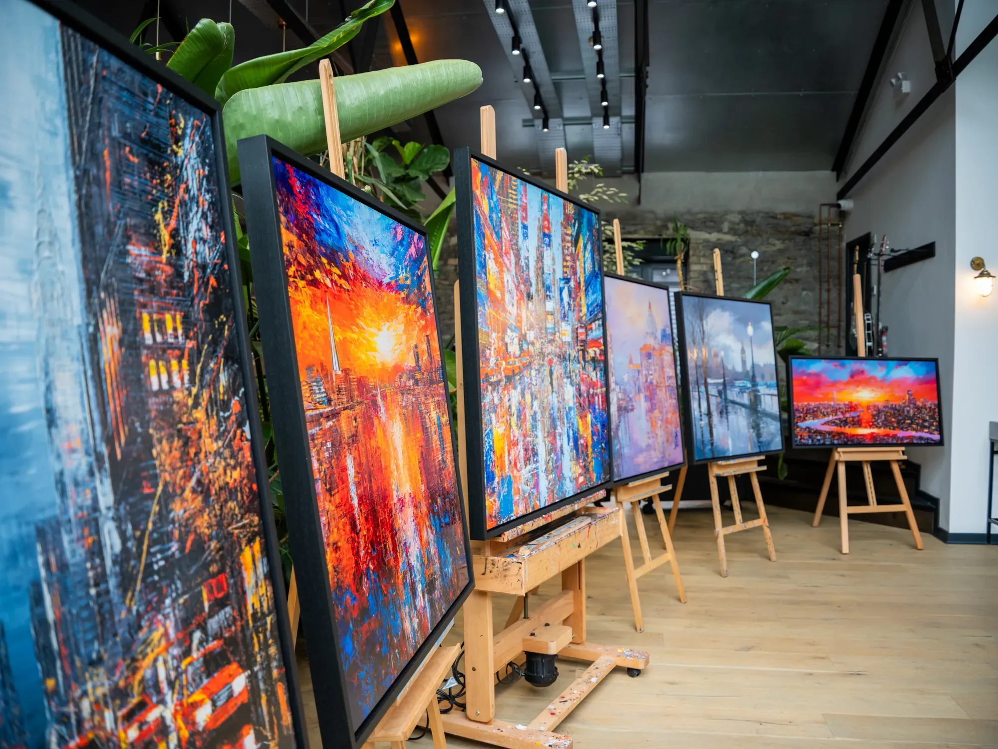 A row of Paul Kenton's colourful cityscape paintings on wooden easels in a bright room with a stone wall and large plants