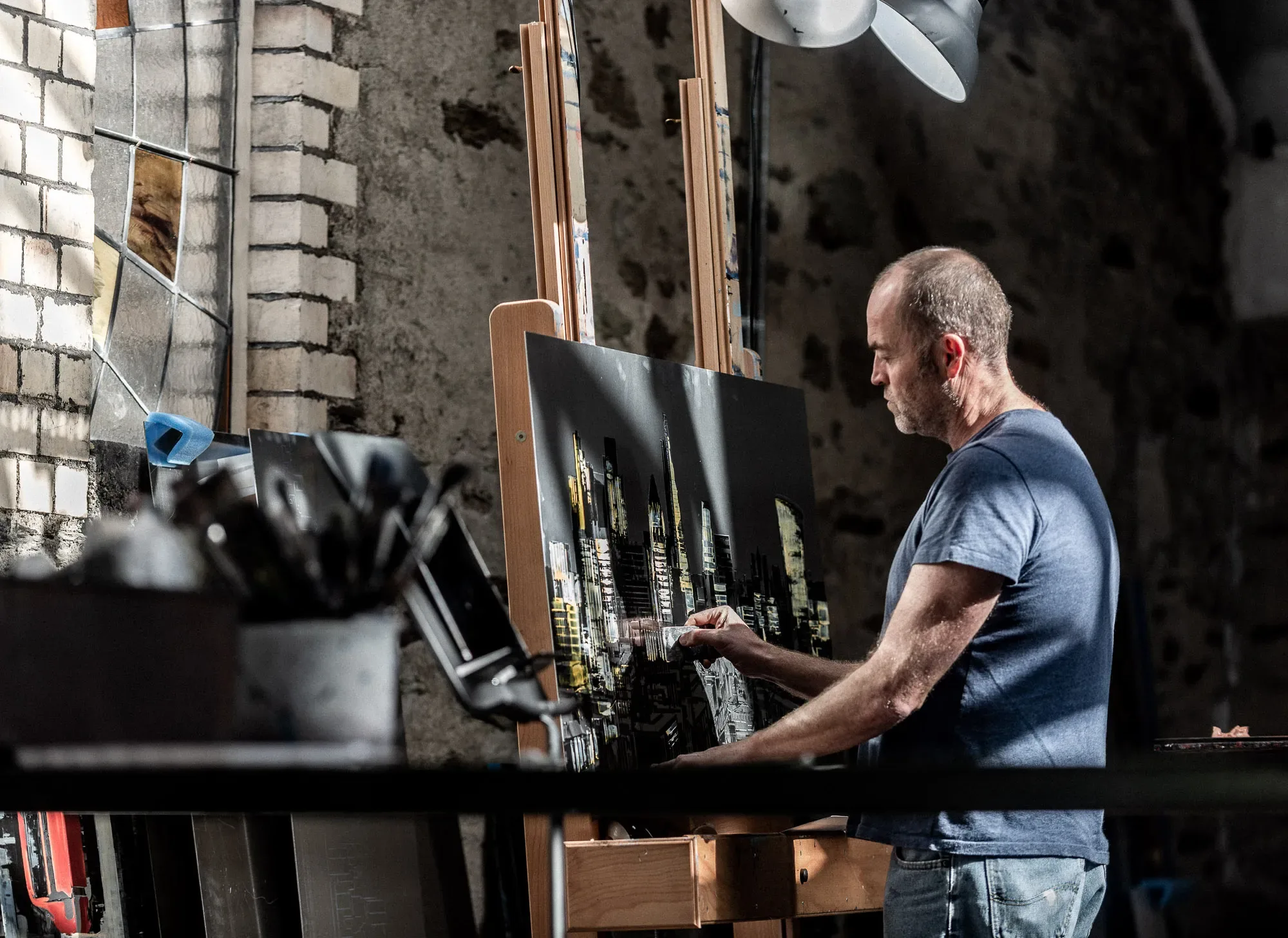 Paul Kenton in a blue T-shirt painting a city skyline at night on an easel, in sunlight against a stone wall