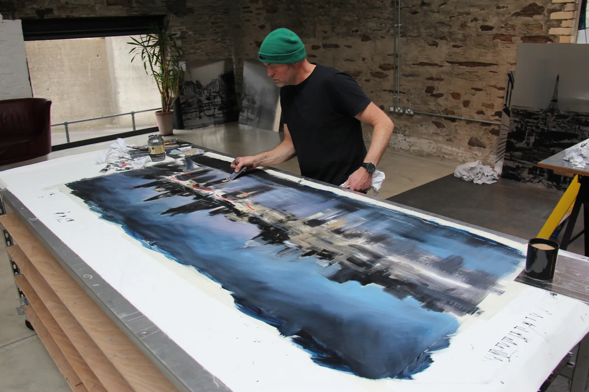 Paul Kenton leaning over a large blue cityscape canvas laid flat on a long table, working into it