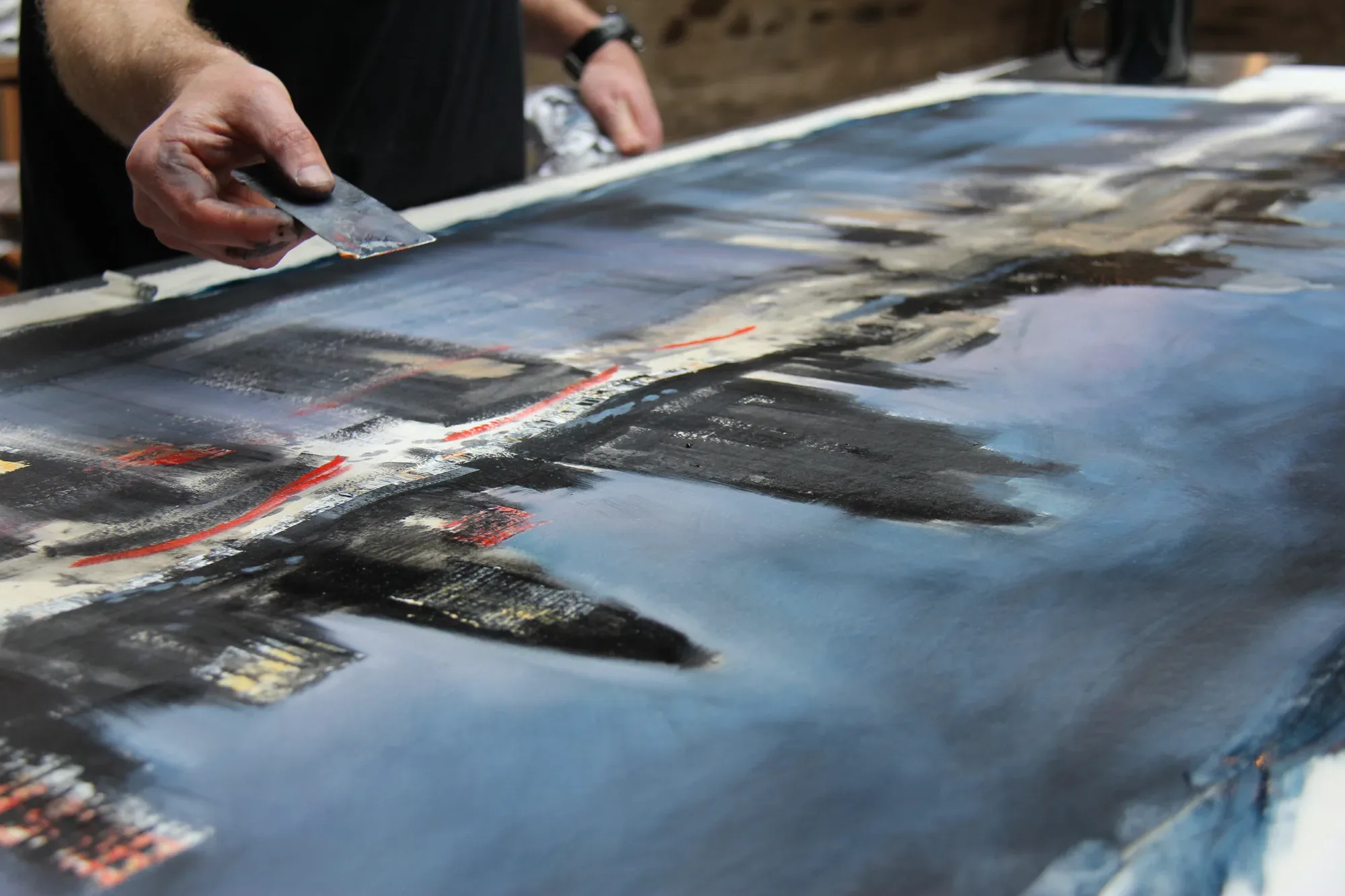 Close-up of Paul Kenton's hand dragging a metal scraper across a dark blue cityscape painting