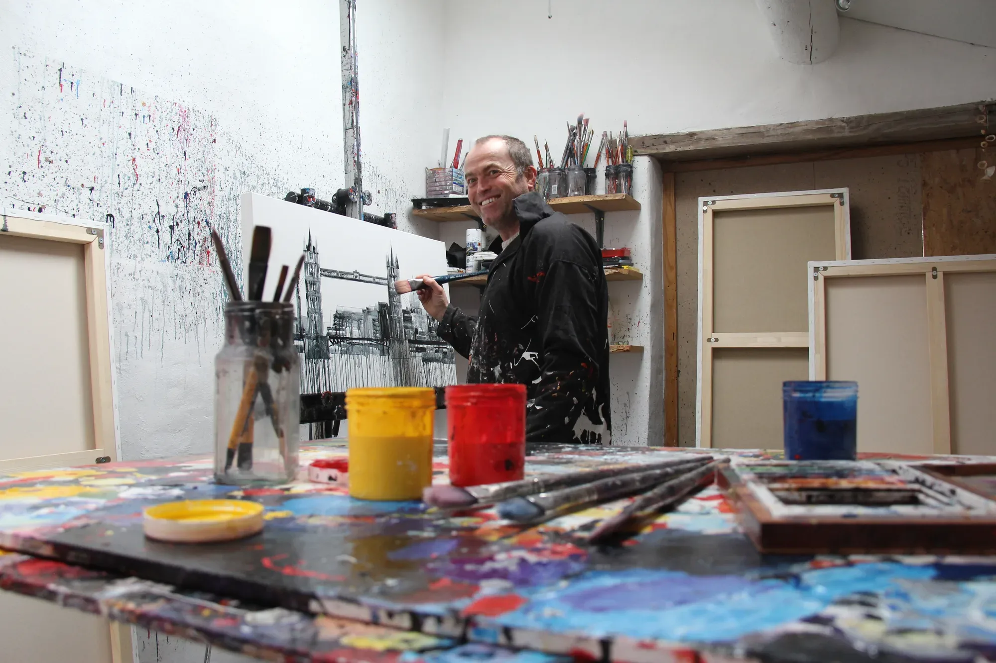 Paul Kenton smiling at the camera as he paints a black and white Tower Bridge canvas, a paint-splashed table in front of him
