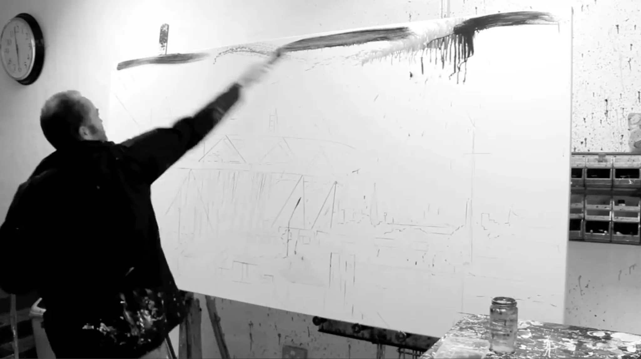 Black-and-white still of Paul Kenton in the studio sweeping a long brush of black paint across the top of a huge canvas, the stadium sketched beneath