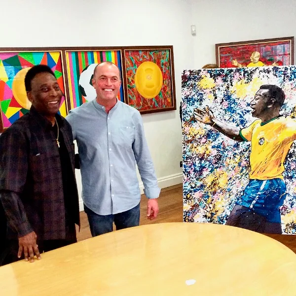 Pelé and Paul Kenton smiling side by side in a gallery, beside Paul’s painting of Pelé celebrating, with other artworks on the wall behind