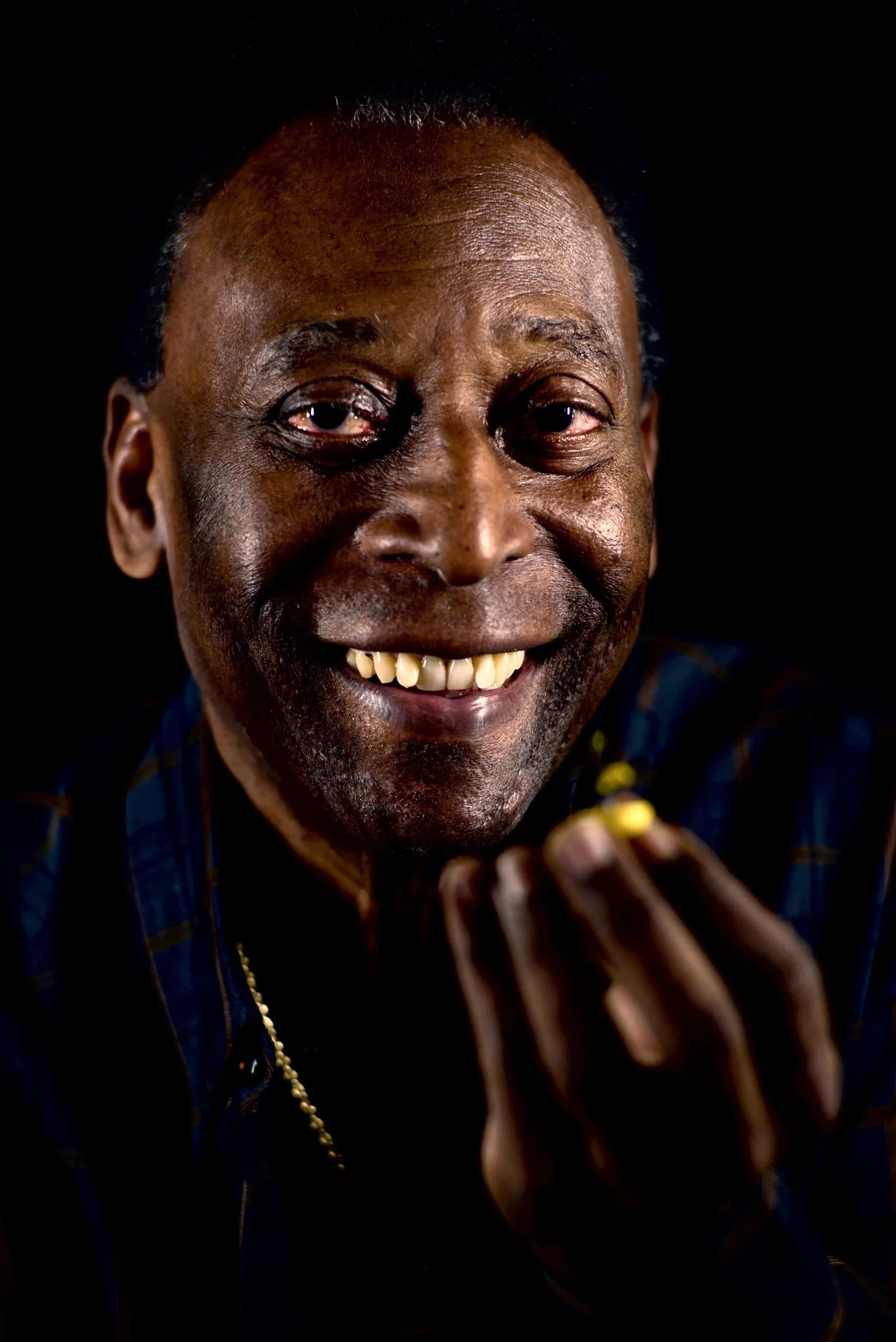 Close portrait of Pelé laughing against a black background, one hand raised towards the camera