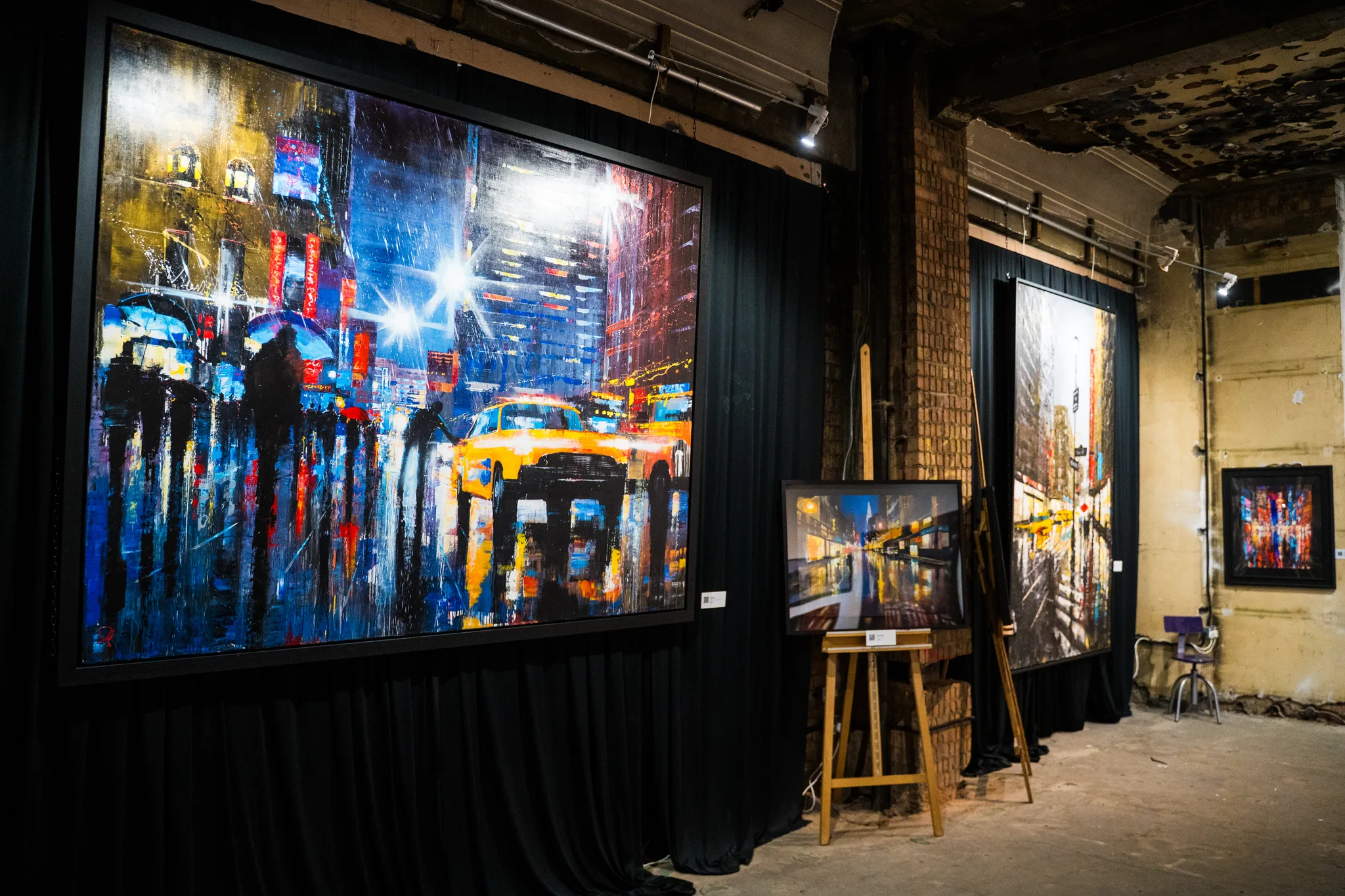 A huge rainy-night street painting, with a yellow taxi and dark umbrellas, hangs on black drapes beside a small easel and a smaller canvas.