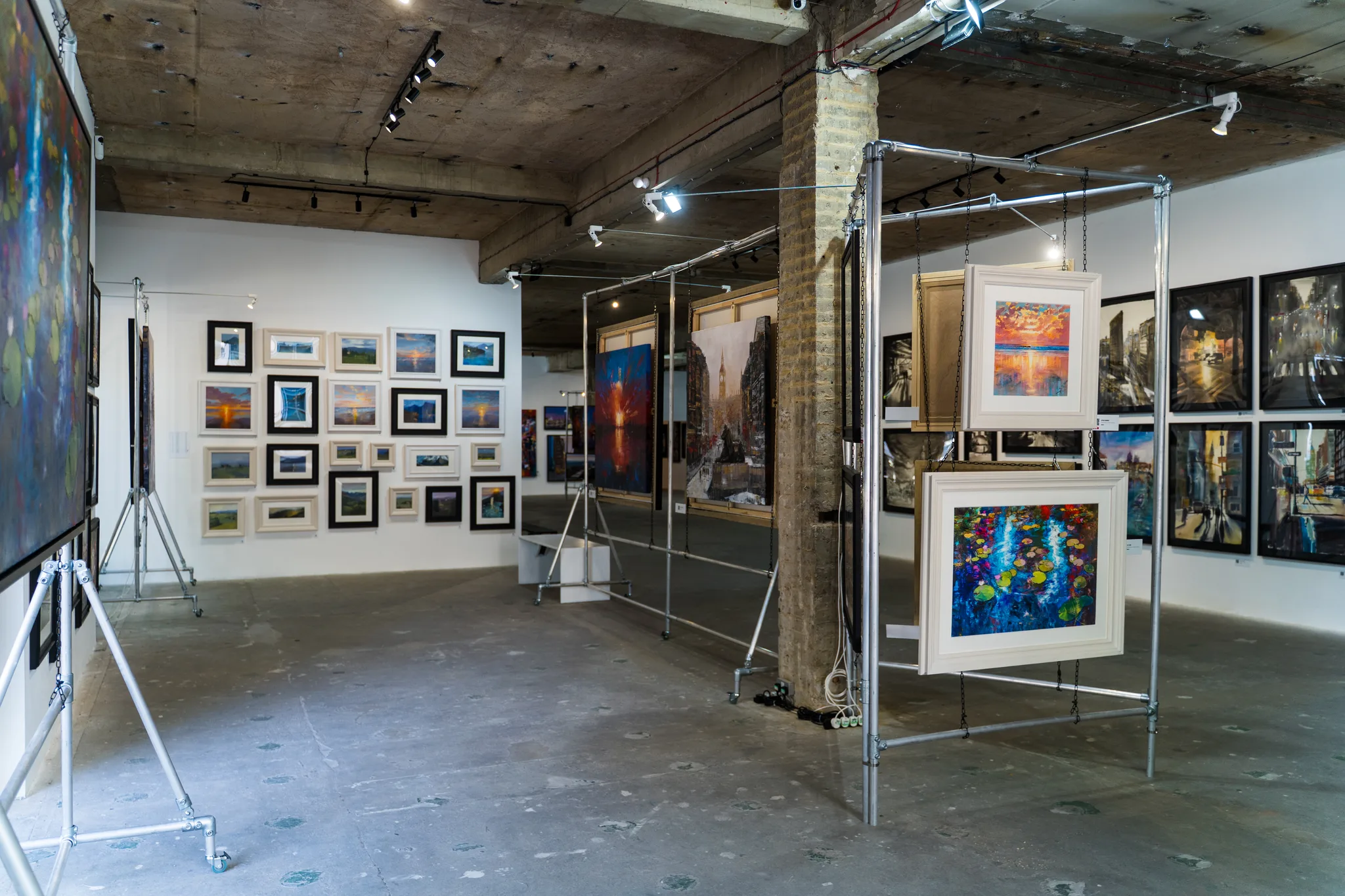 Small framed works cover a white wall, while a steel frame in the centre holds a larger picture of bright blue and red shapes in a pale frame.