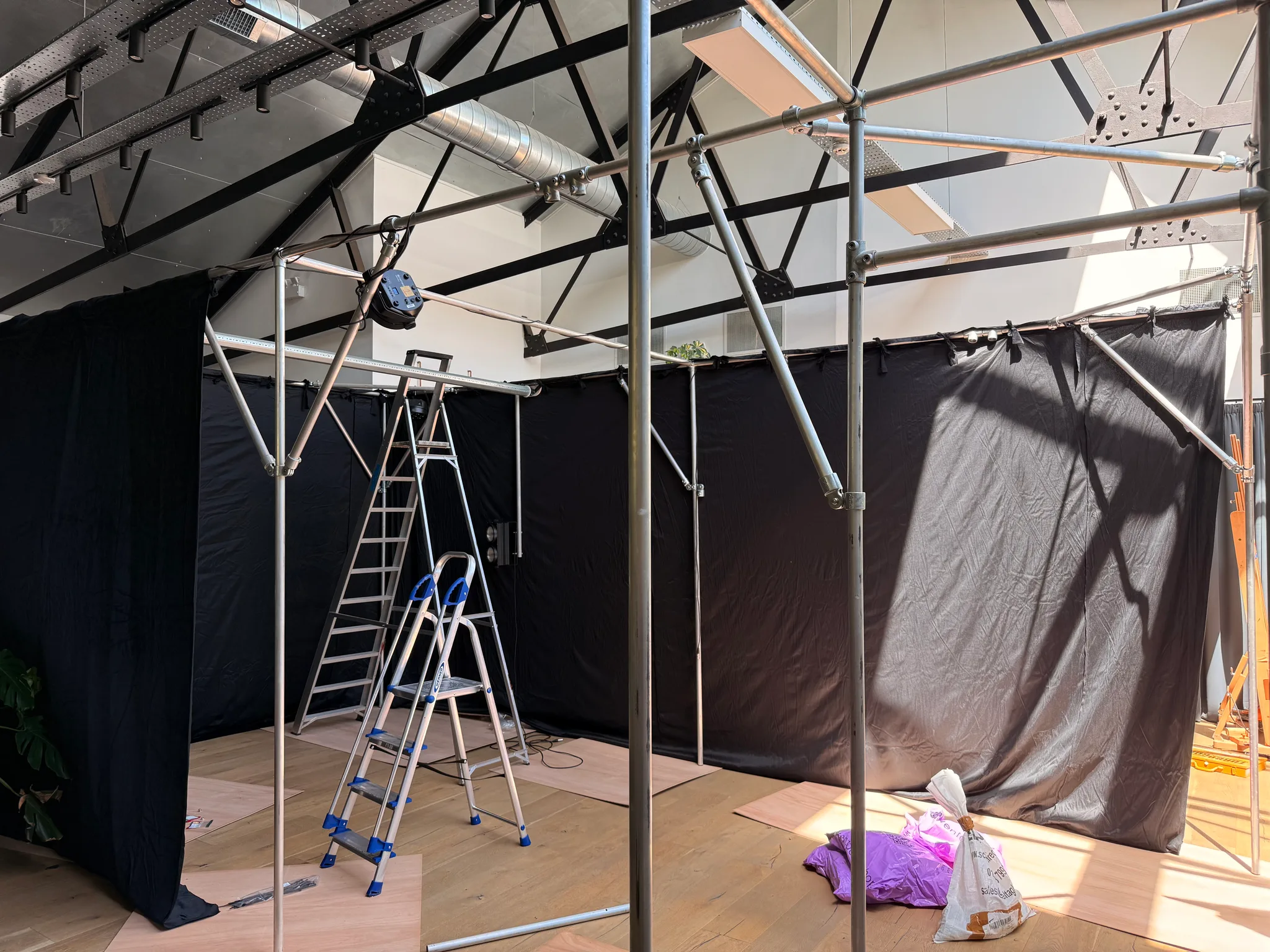 Black drapes hang from a steel scaffold in a sunlit room with a timber floor, where a stepladder and a tall ladder stand amid the drapes.