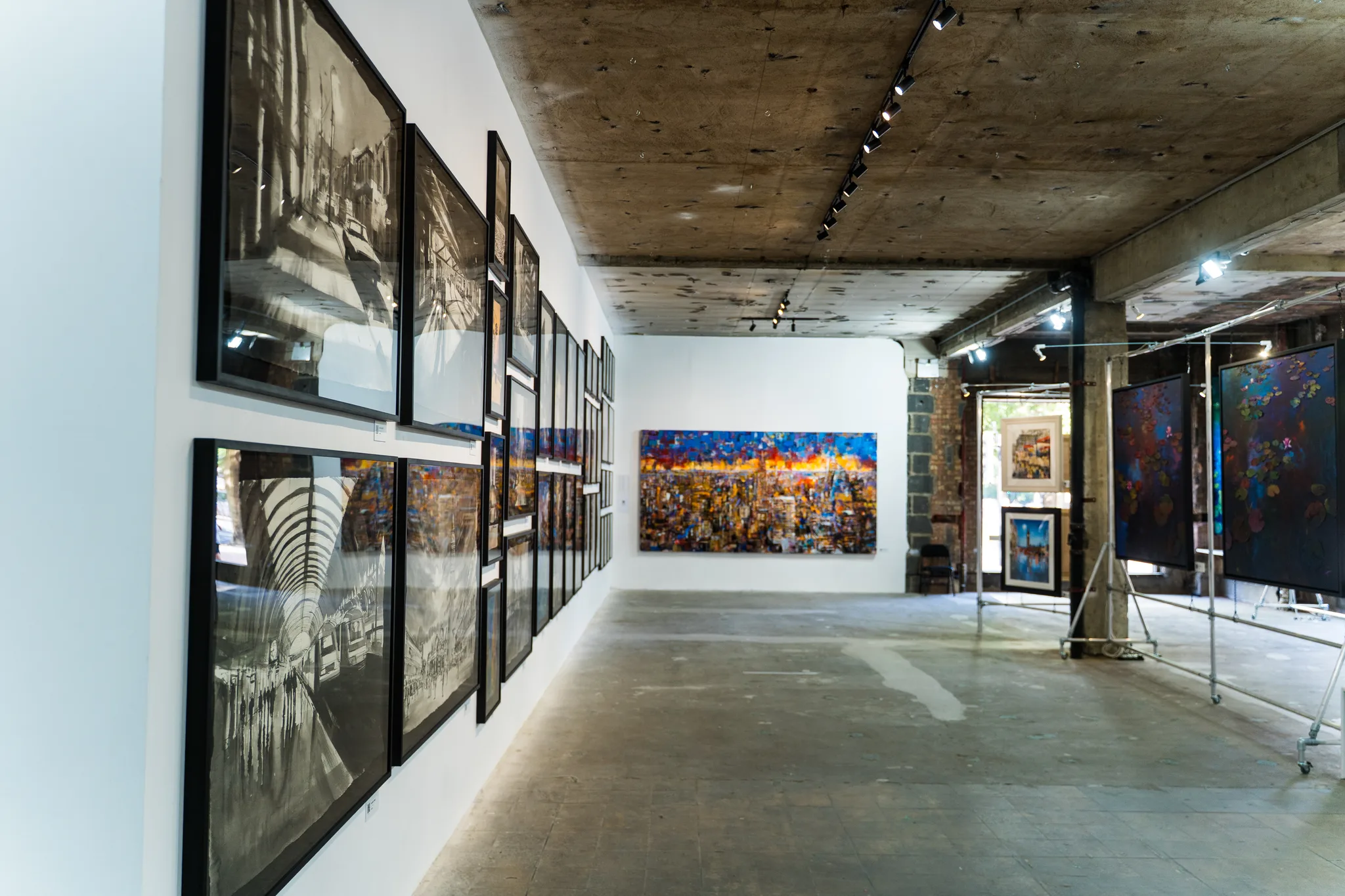 A long concrete-floored room with black-and-white city drawings in black frames lining the left wall and a bright city painting at the far end.