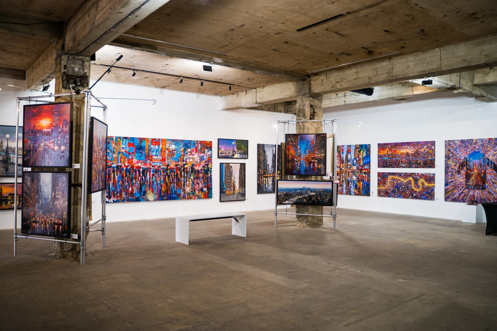 A raw concrete-beamed hall with white walls, where bright city paintings hang on steel frames and a white bench sits on the grey floor.
