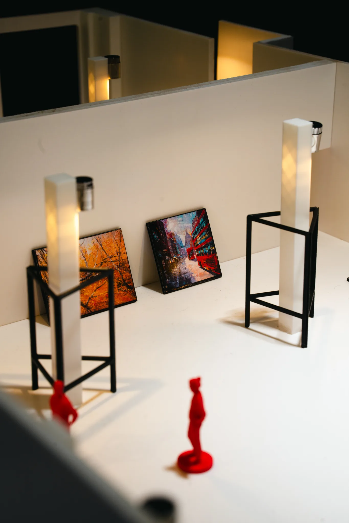 Close view of a white model gallery, with two tiny paintings on black wire stands beside glowing pillars and two red figures in the foreground.