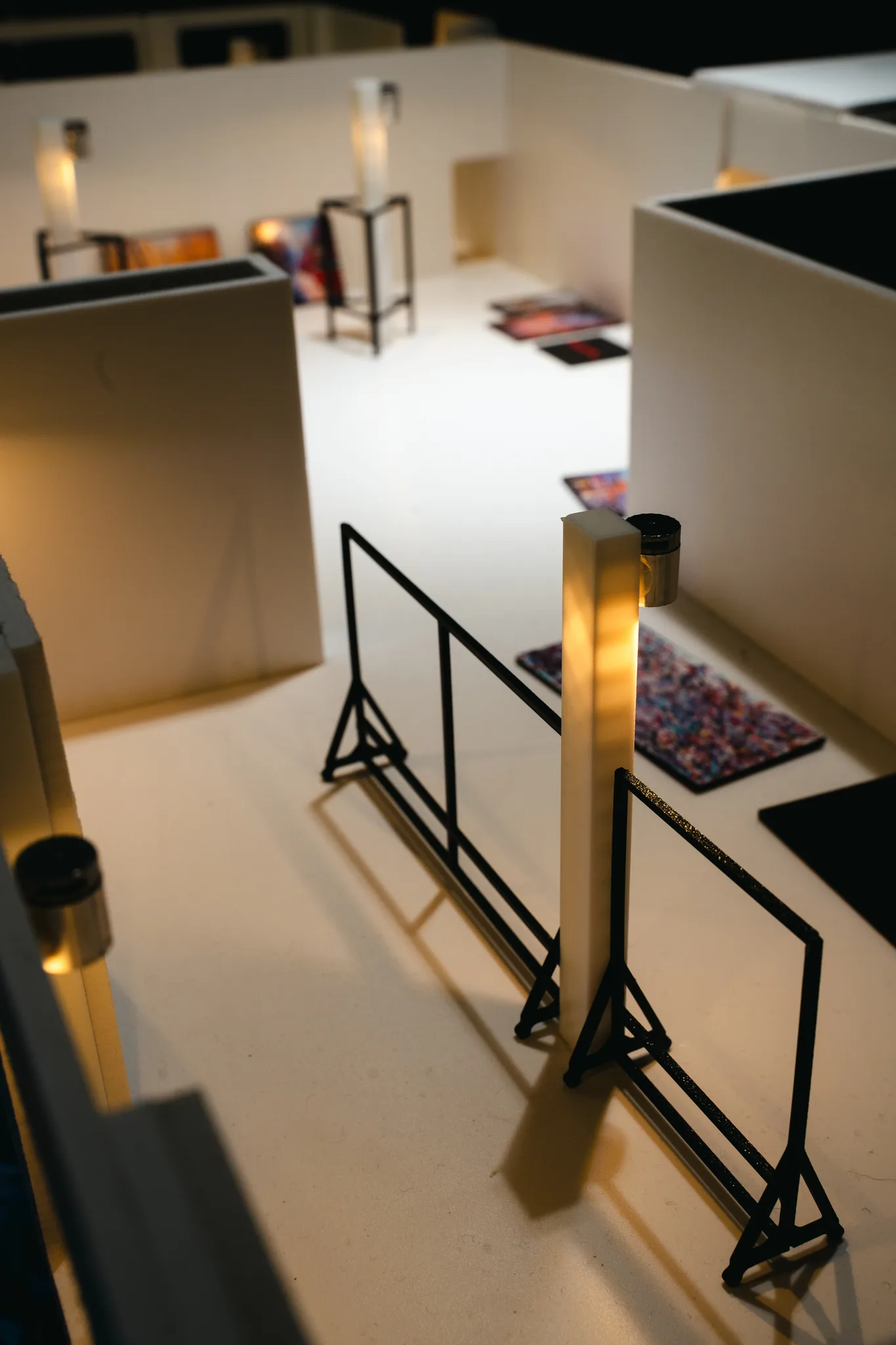 Looking down a model gallery corridor past a black wire railing and glowing white pillars, with tiny paintings lying on the floor beyond.