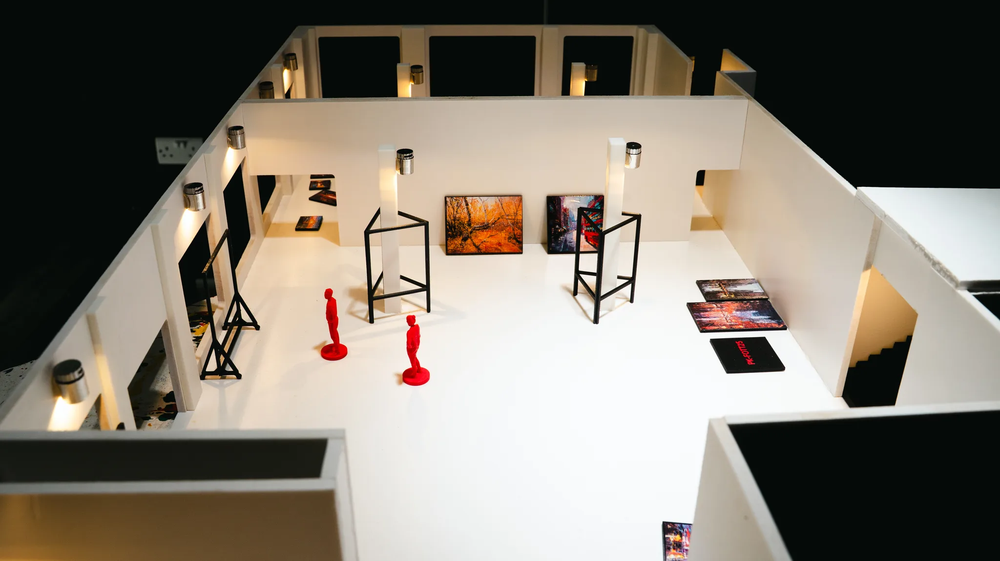 Overhead view of the white model gallery: two red figures, black wire stands, and tiny paintings on the floor, with the dark model walls around it.