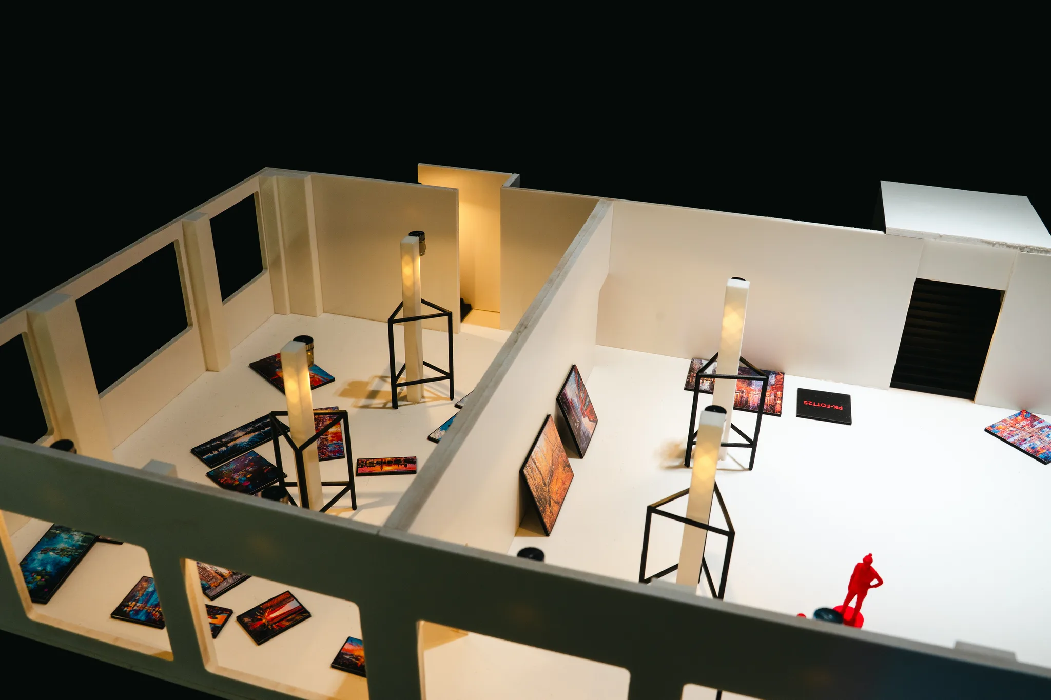 Elevated view across the model’s rooms, with tiny paintings scattered over the white floor, black wire stands by glowing pillars and one red figure at right.
