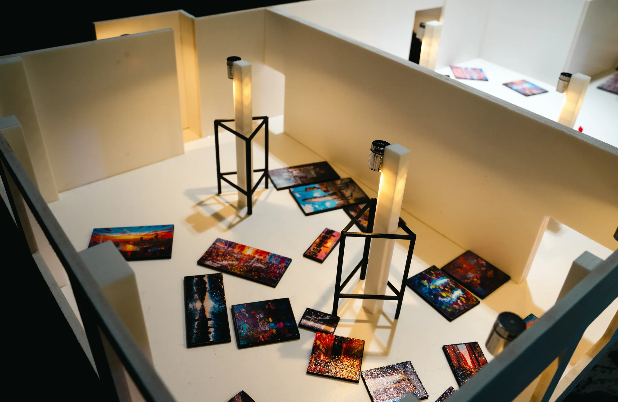 Angled close view of the model floor, with tiny city paintings spread around black wire stands and a glowing white pillar.