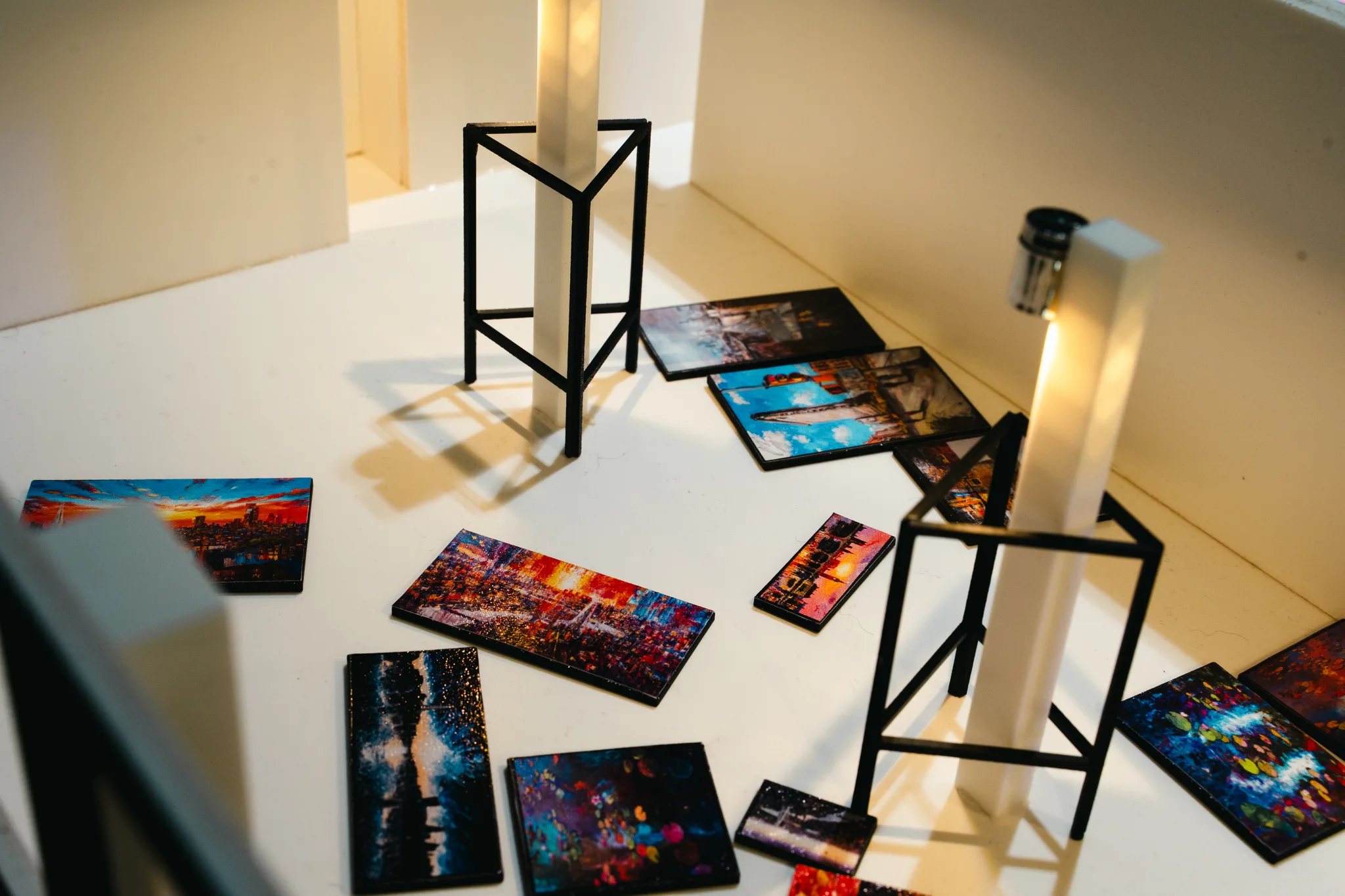 Tiny paintings lie scattered across the white model floor beside glowing pillars and black wire stands, under warm, low light.