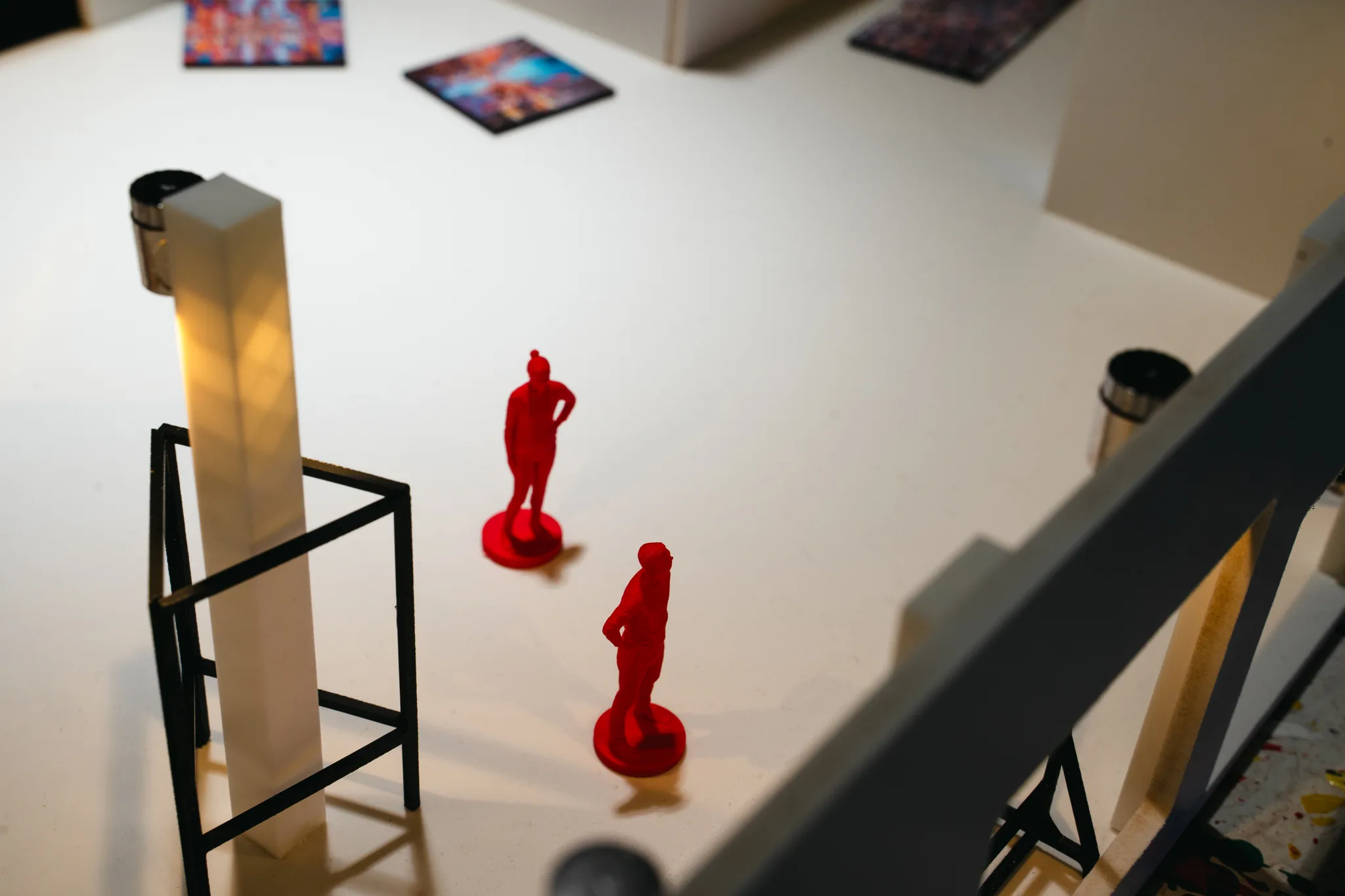 Two red model figures stand on the white floor among tiny paintings, beside a glowing pillar and a black wire stand.