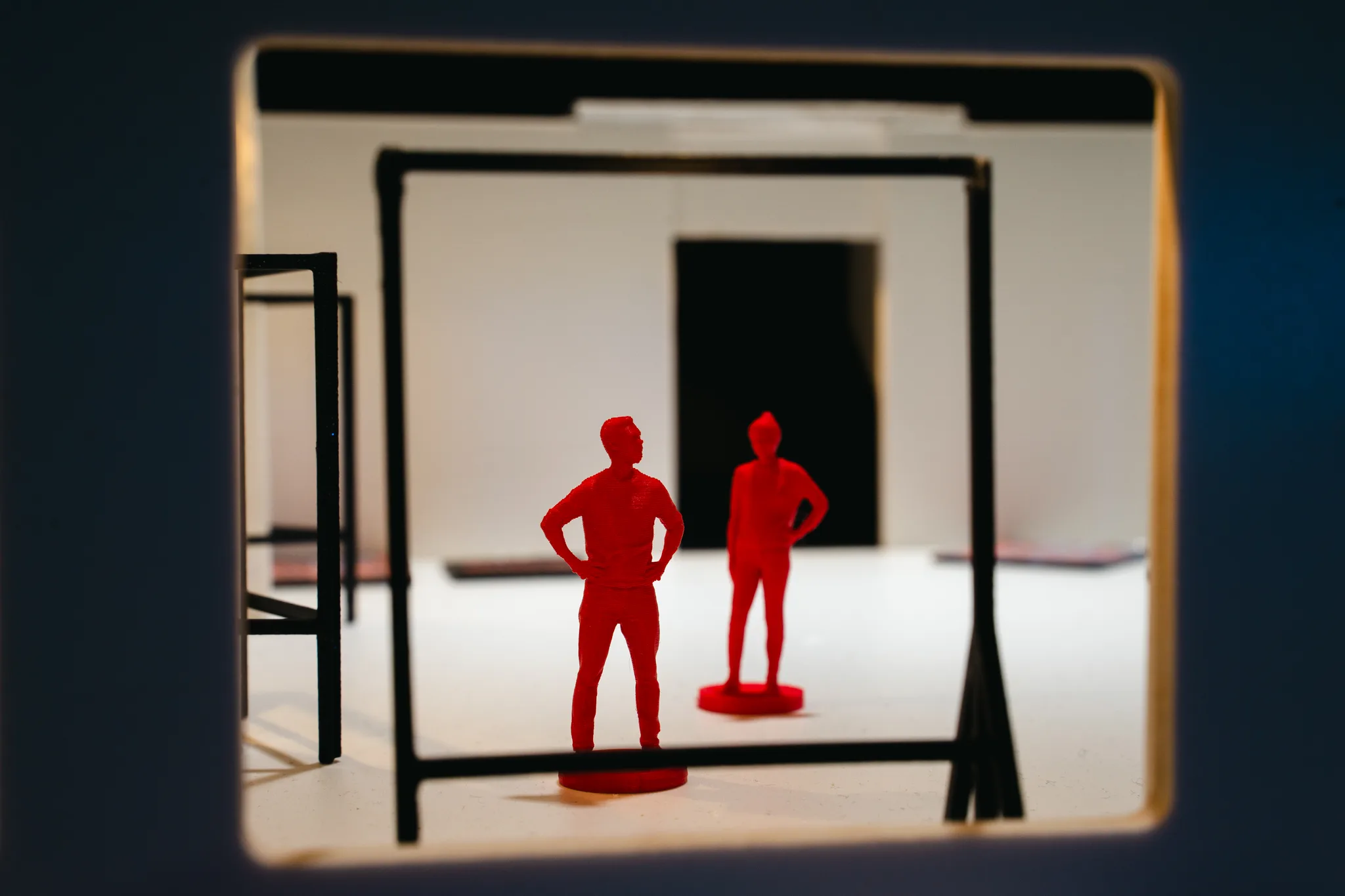 Seen through a model doorway, two red figures stand inside a black wire frame, with a white gallery room and dark opening beyond.