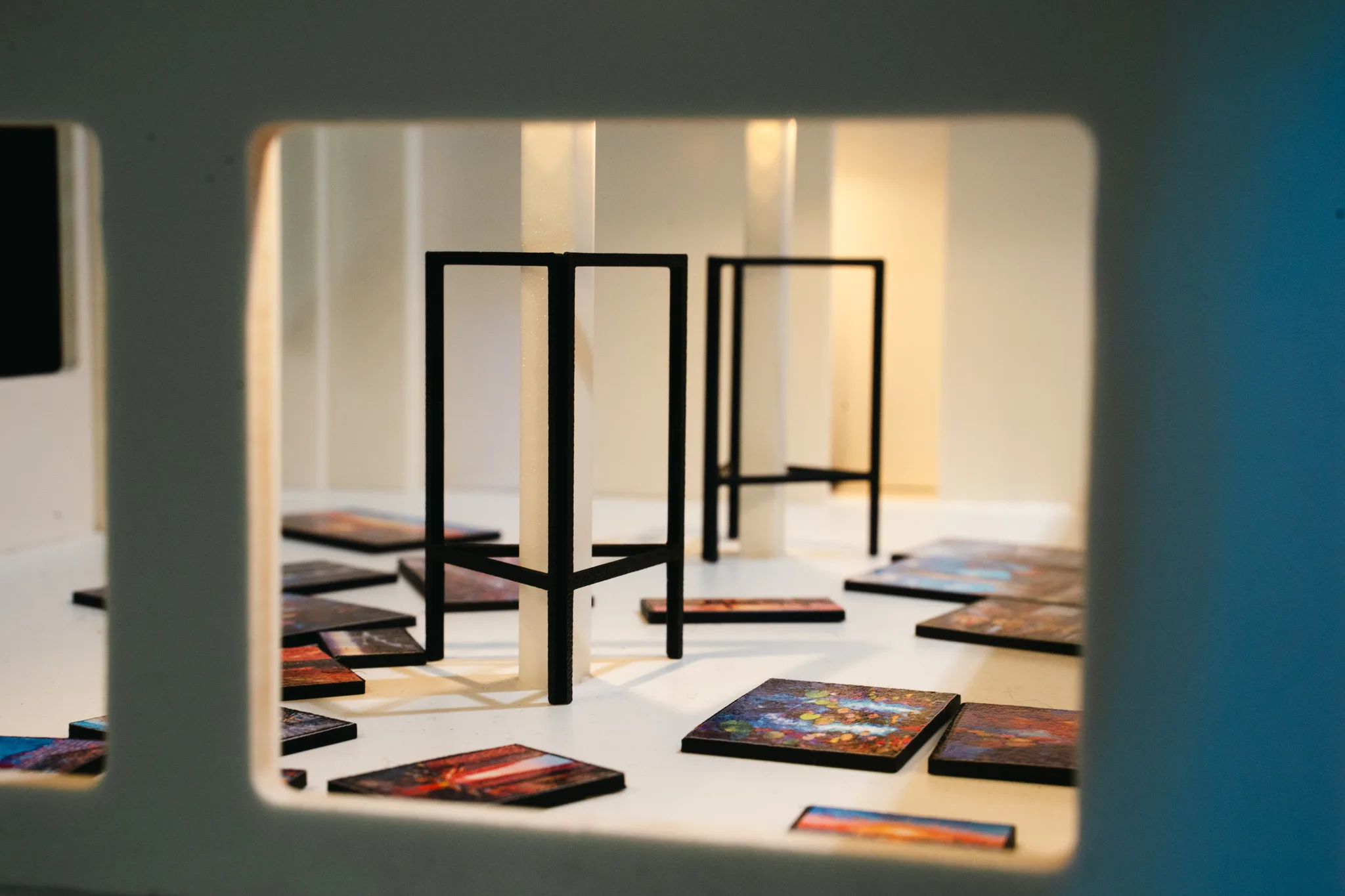 Through a window cut in a model wall, black wire stands and tiny paintings on the white floor sit in soft light beside glowing pillars.