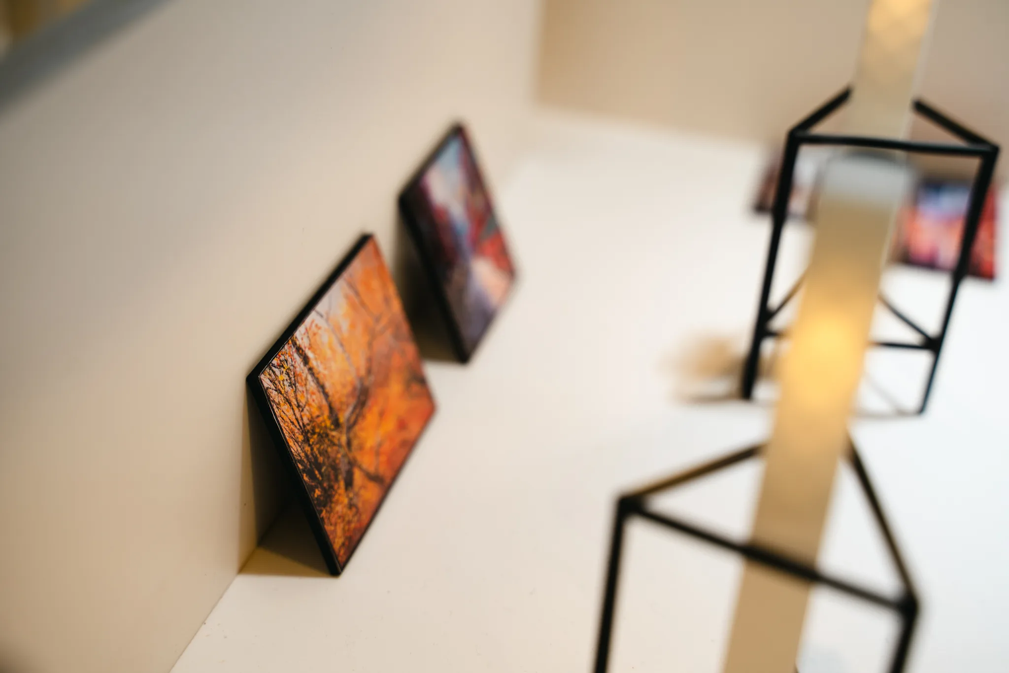 Two tiny paintings lean against a cream model wall, orange and purple, with blurred black wire stands and a glowing pillar behind.