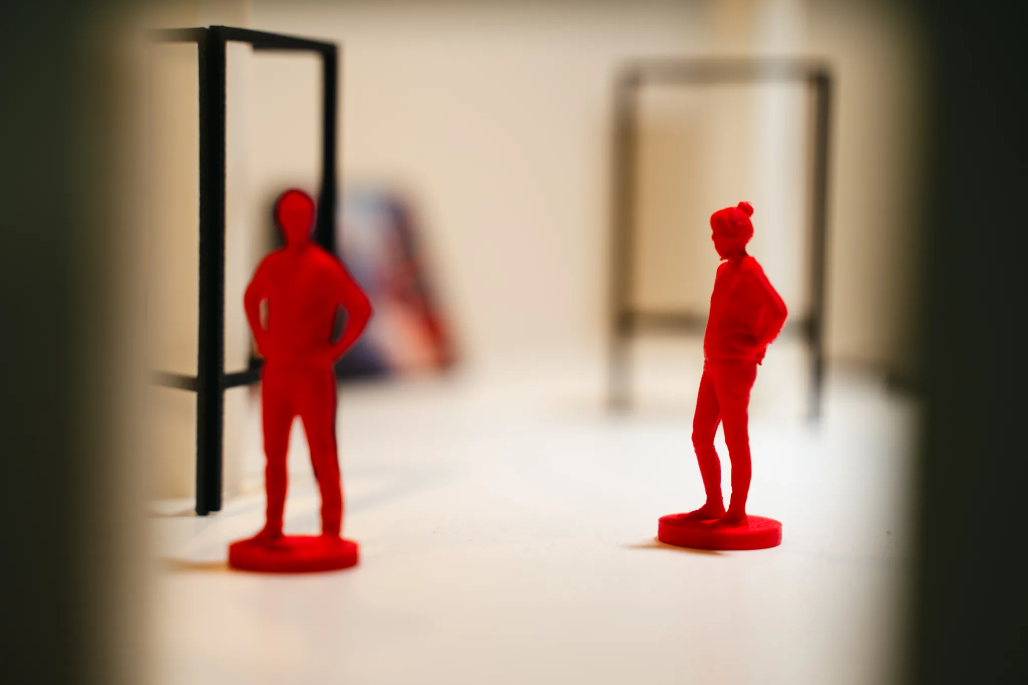Two red model figures stand on the white floor beside a black wire stand, with a blurred painting in the background.