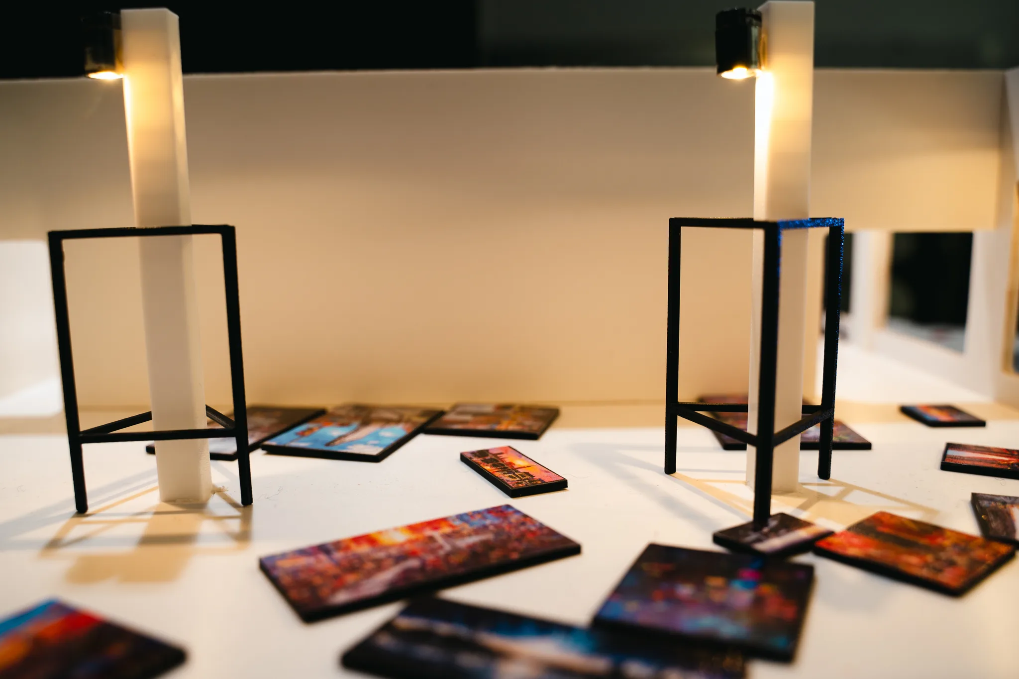 Tiny paintings lie across the warm-lit model floor, with black wire stands and glowing pillars on either side.
