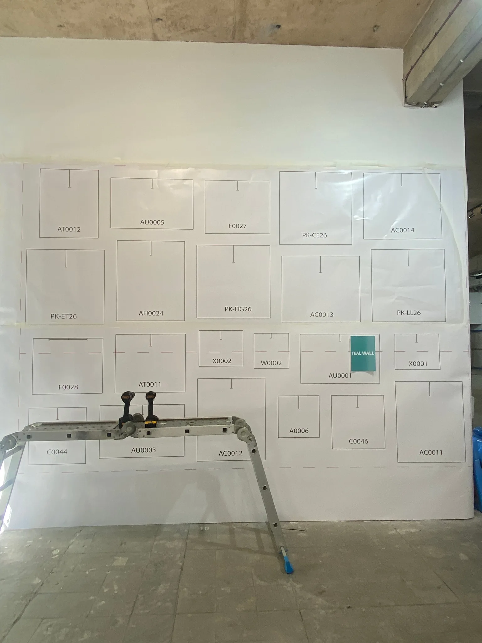 A white wall covered in paper outlines of paintings, each labelled with a code such as PK-CE26, with a teal square and a folding stepladder in front.