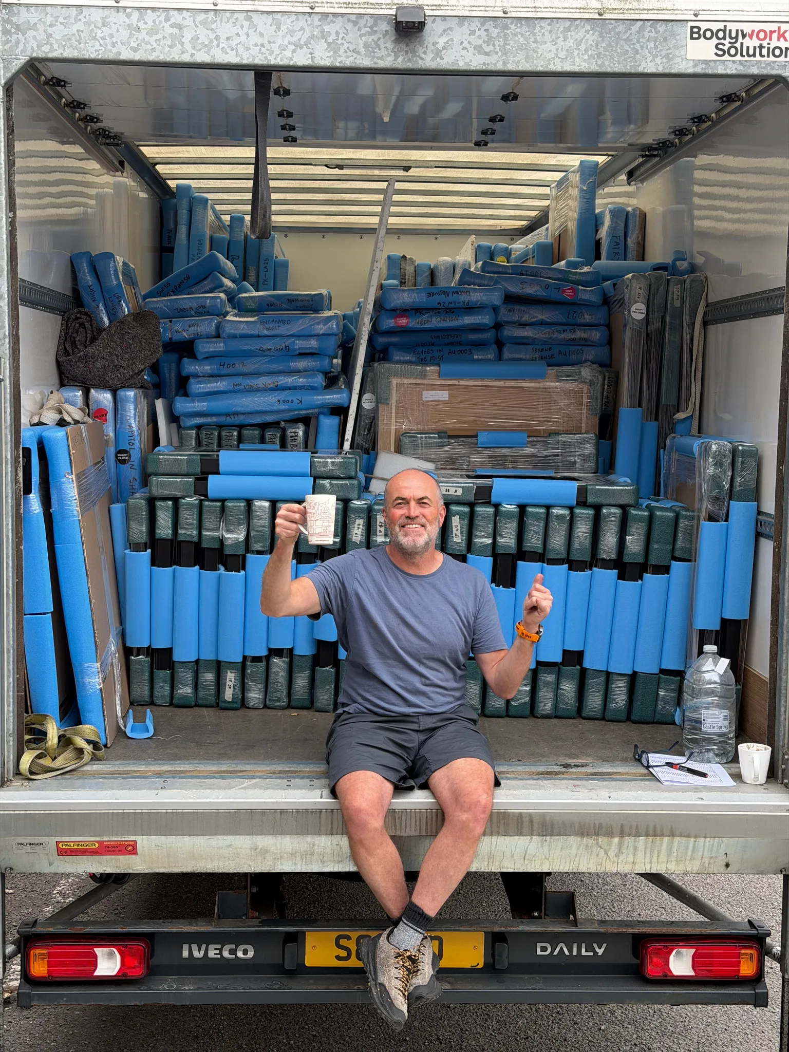 Paul Kenton sits on the tail lift of a van stacked high with blue-wrapped paintings, raising a mug in one hand and a fist in the other.