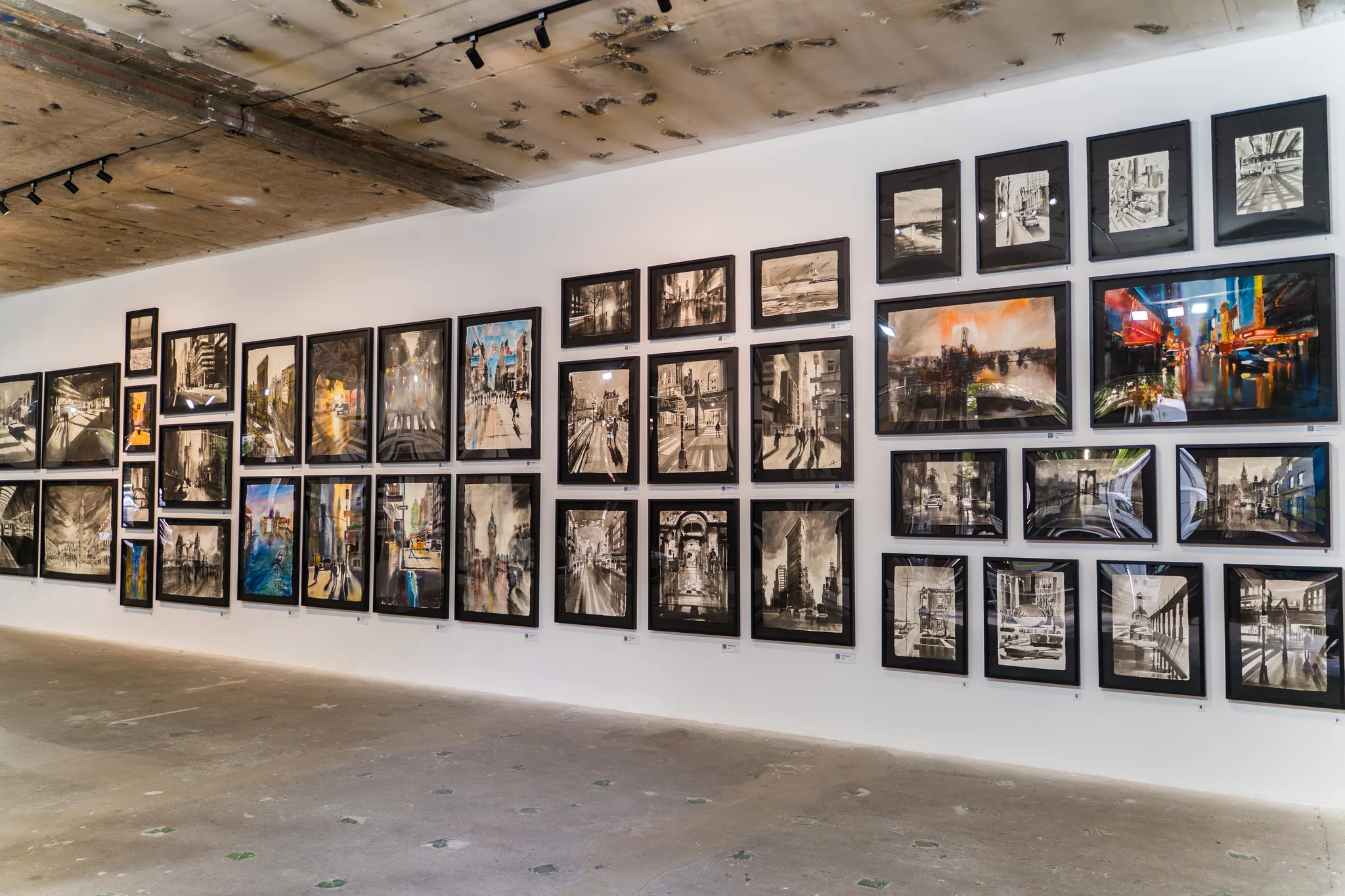 A crowded salon-style wall of dozens of black-framed city pictures, mostly black-and-white, with a few colour paintings, under a concrete ceiling.