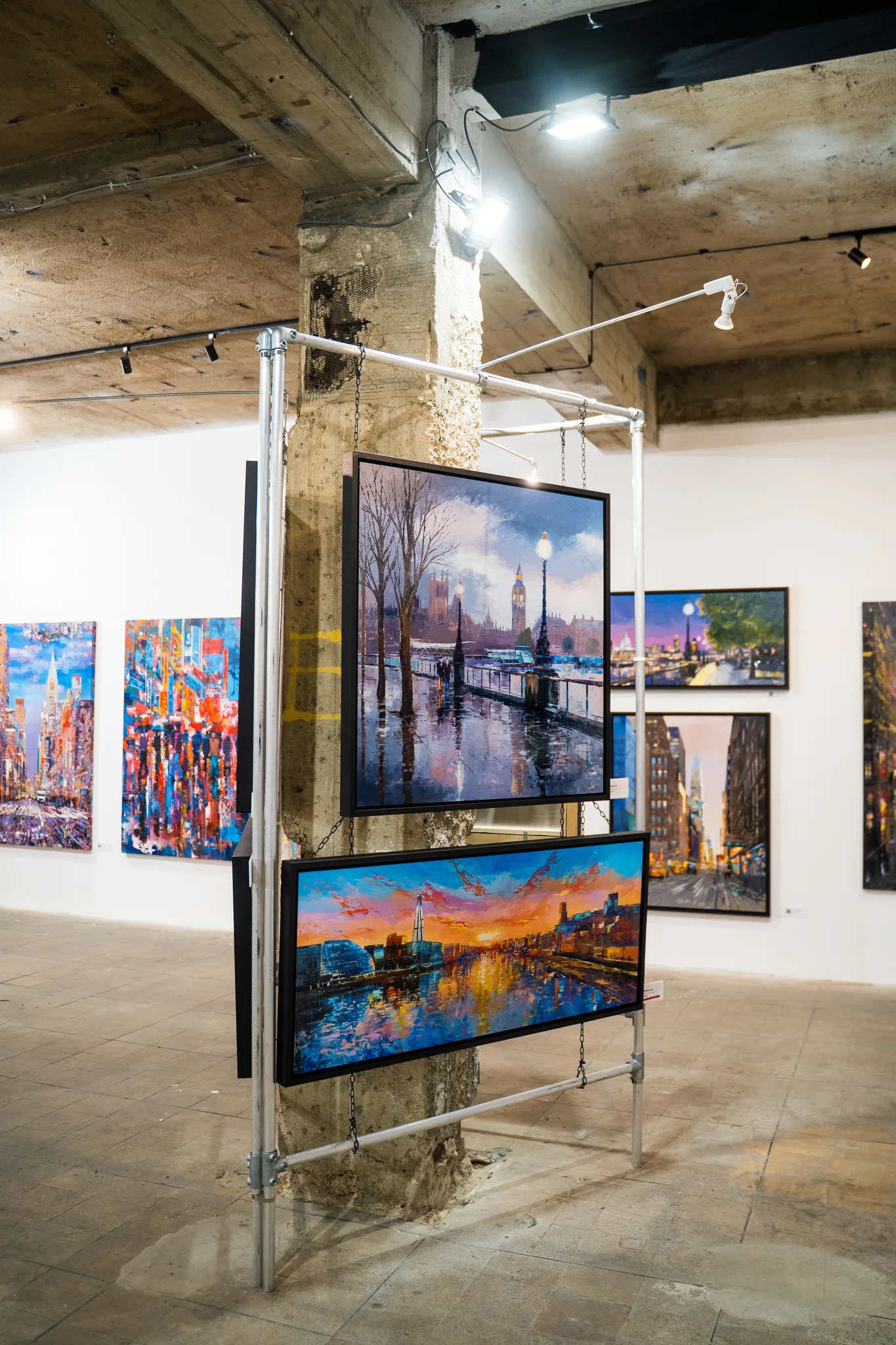 Two paintings hang on a steel frame beside a rough concrete column: a misty Westminster riverside scene above a glowing orange sunset over water.