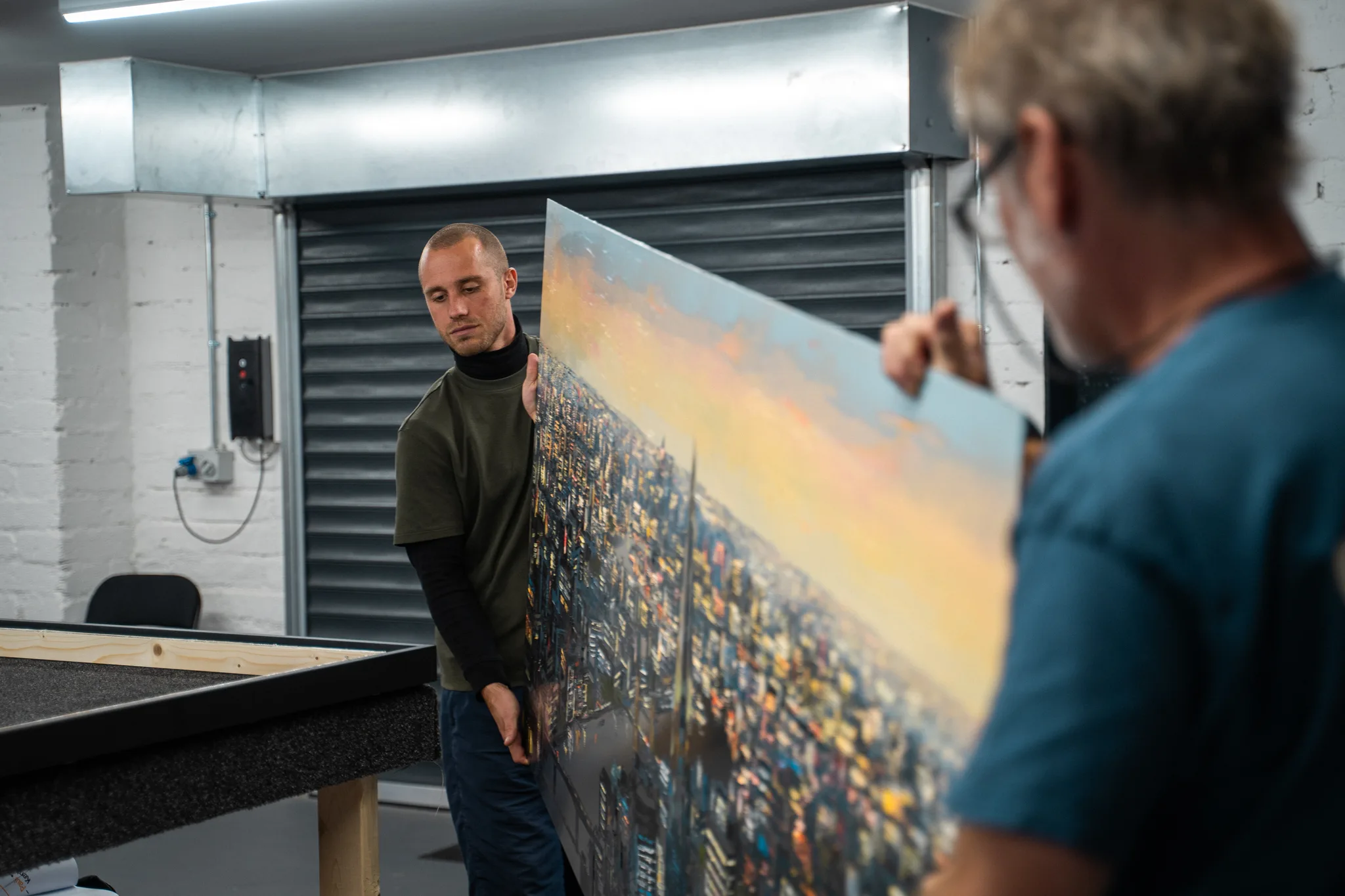Alan and a colleague holding a large cityscape on aluminium upright in the framing workshop