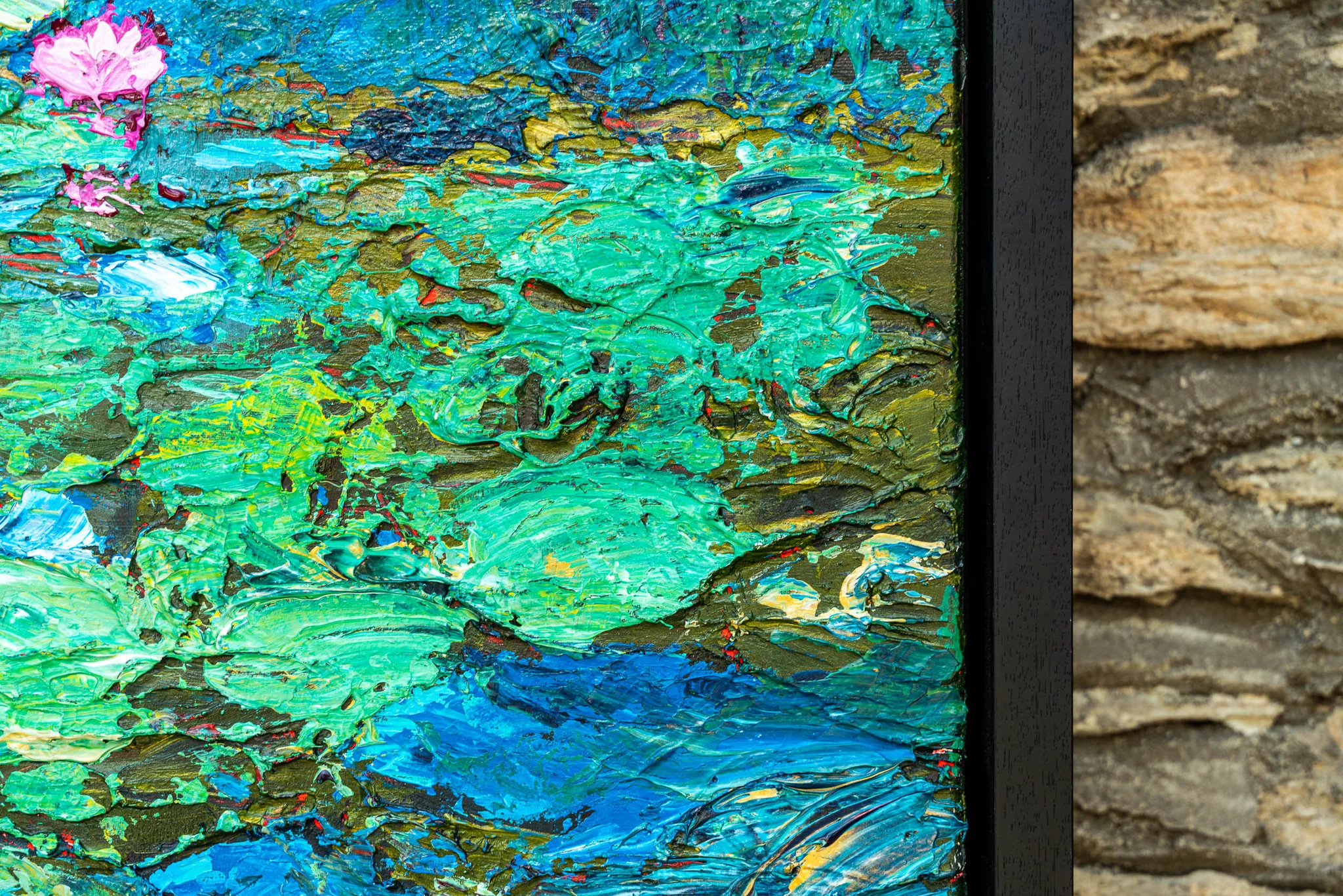 Close-up of Impressionist Waters, an original painting by Paul Kenton: thick, textured strokes of green, turquoise and blue with a small pink water lily, set in a black frame beside a stone wall.