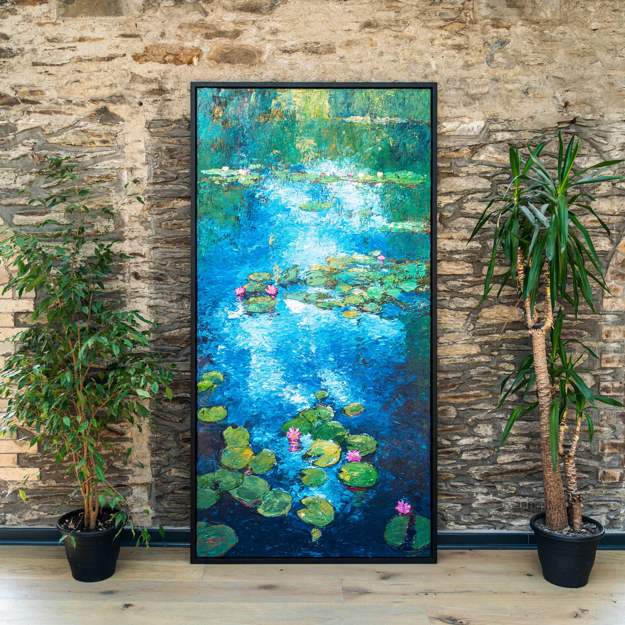 Impressionist Waters, an original painting by Paul Kenton, leans against a rough stone wall between two potted plants, showing a textured pond of blue water, green lily pads and pink water lilies.