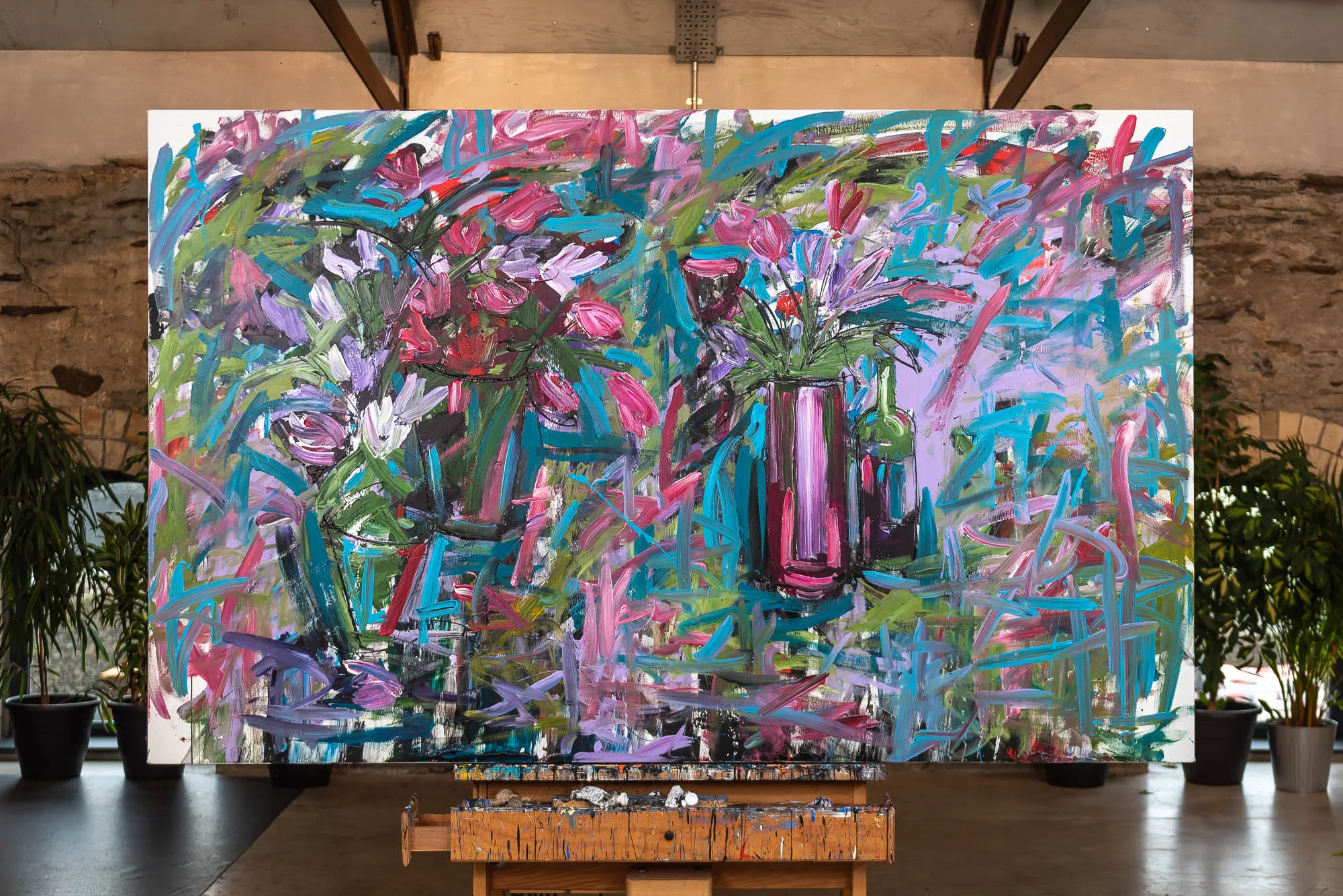 Joy, an original mixed media painting by Paul Kenton shown on an easel in a studio: loose, energetic brushstrokes in pink, teal, violet and green form flowers in vases against a bright, lively background.