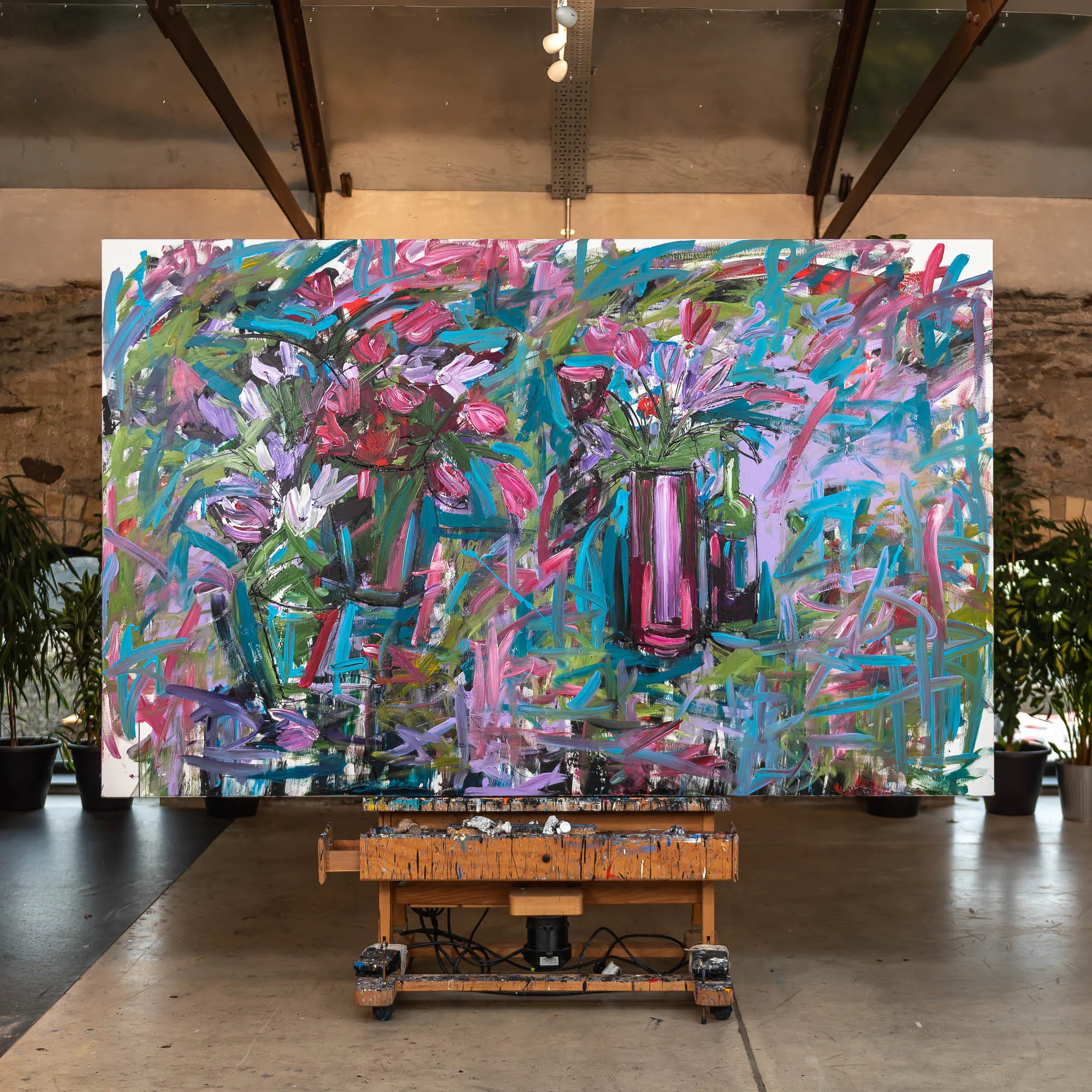 Joy, an original mixed media painting by Paul Kenton, hangs in a large studio on a wooden trestle. Loose, energetic brushstrokes in pink, turquoise and violet fill a canvas of flowers in vases.
