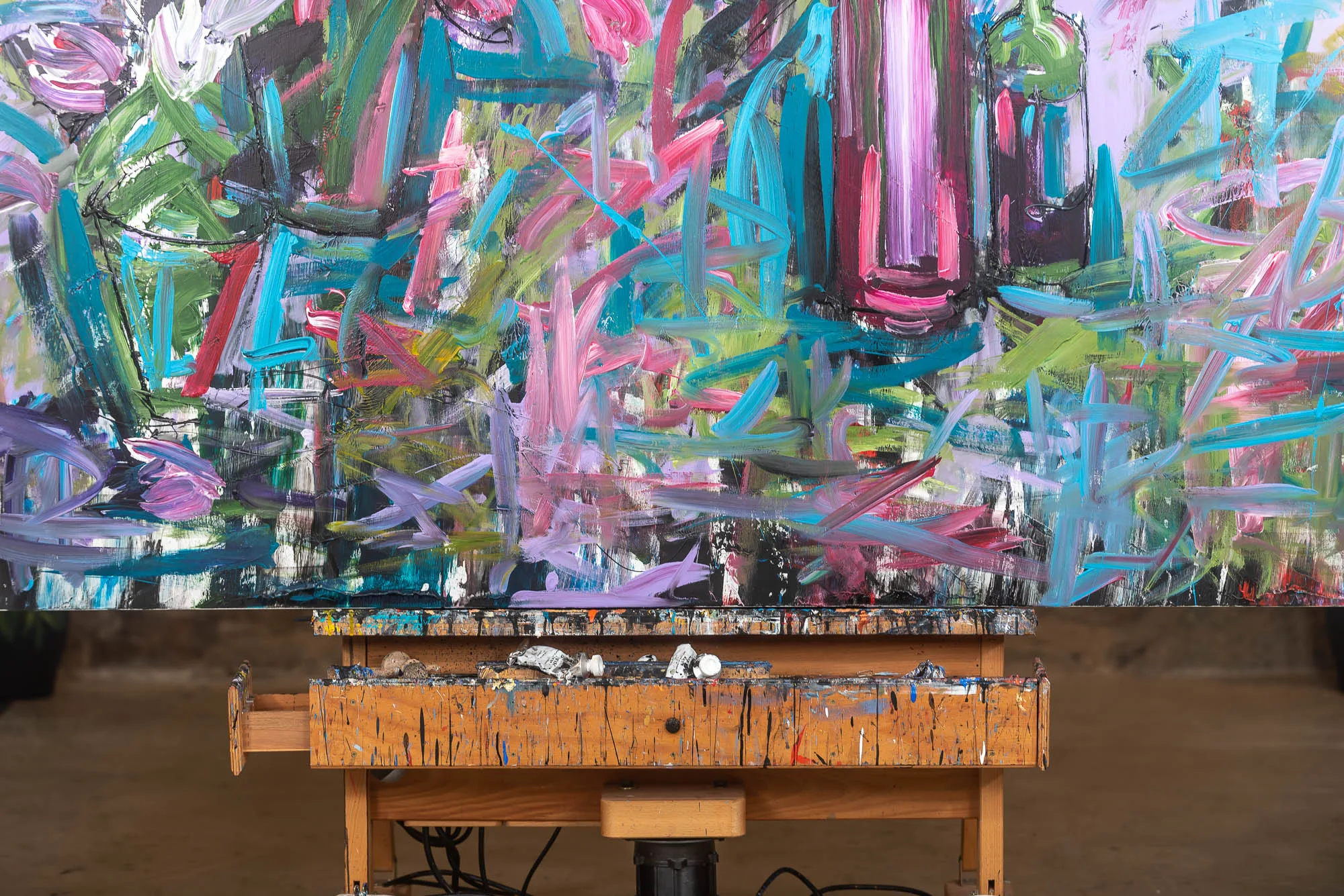 Close-up of Joy, an original mixed media painting by Paul Kenton, showing loose, energetic strokes of turquoise, magenta, green and lilac over a pale ground, resting on a paint-spattered wooden table.