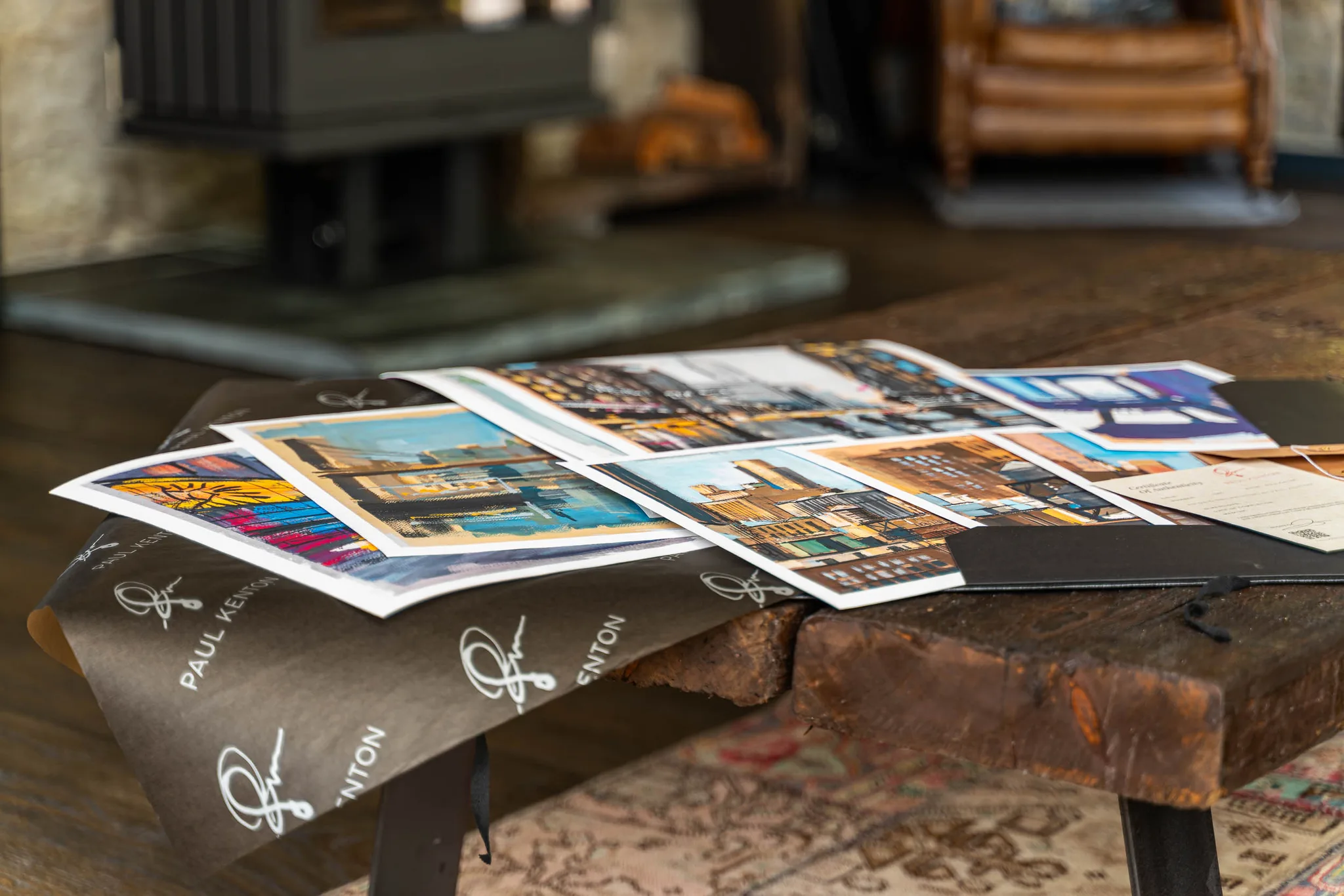 Several Paul Kenton art prints fanned across a wooden table, showing bold, textured New York street scenes in warm oranges, blues and yellows, on paper printed with his signature.