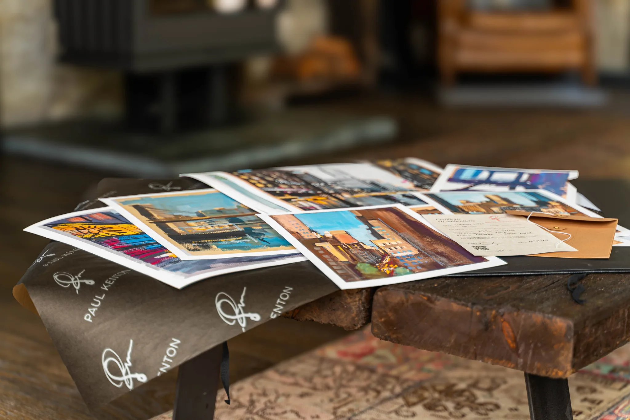 Several Paul Kenton art prints fanned across a wooden table beside a brown wrapping sheet printed with his signature, showing bold, textured New York street scenes in warm oranges and blues.