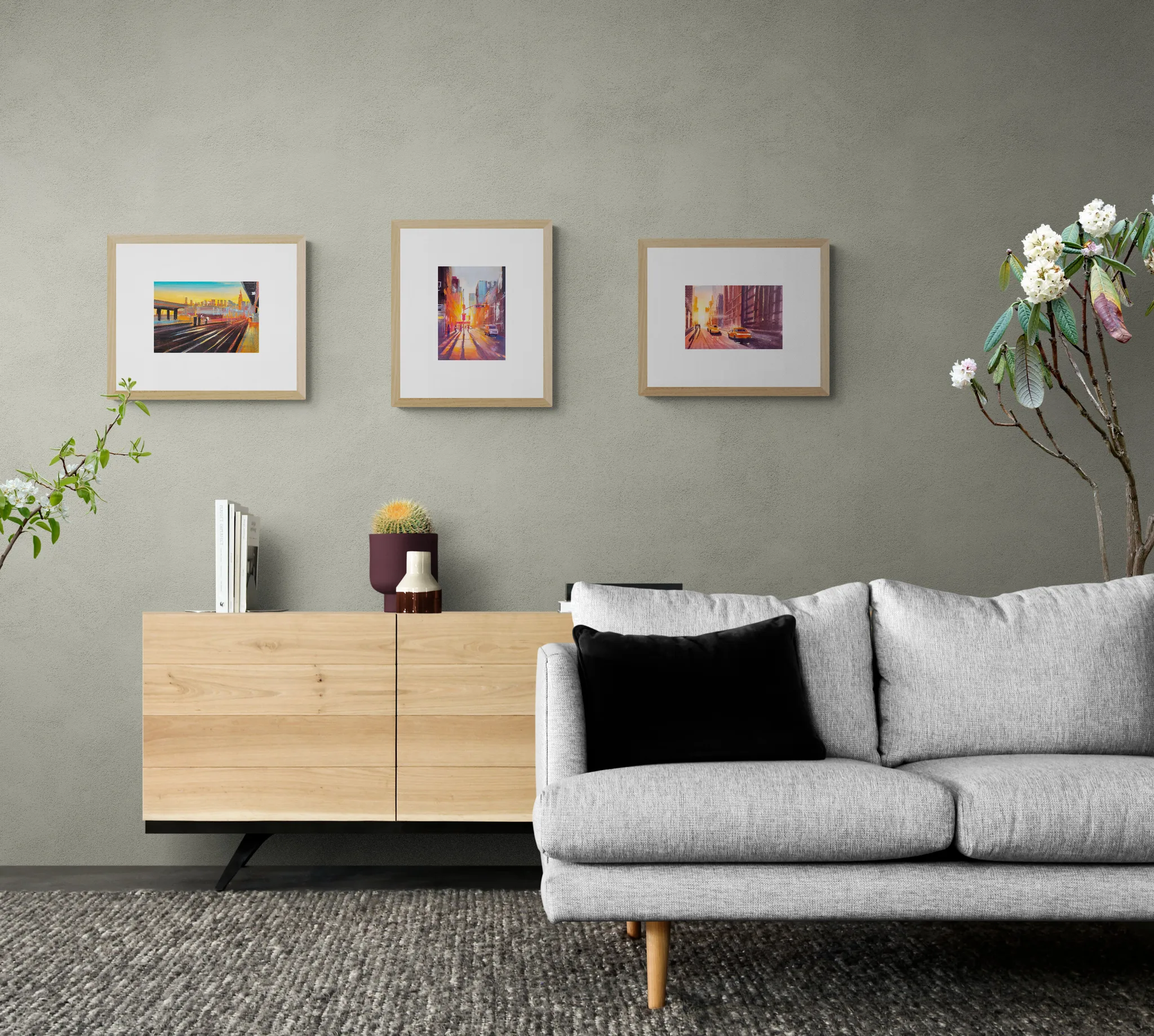Three framed New York Sunsets prints hang above a light oak sideboard in a living room, with a grey sofa and black cushion below; the prints show glowing city street scenes in warm orange and pink.
